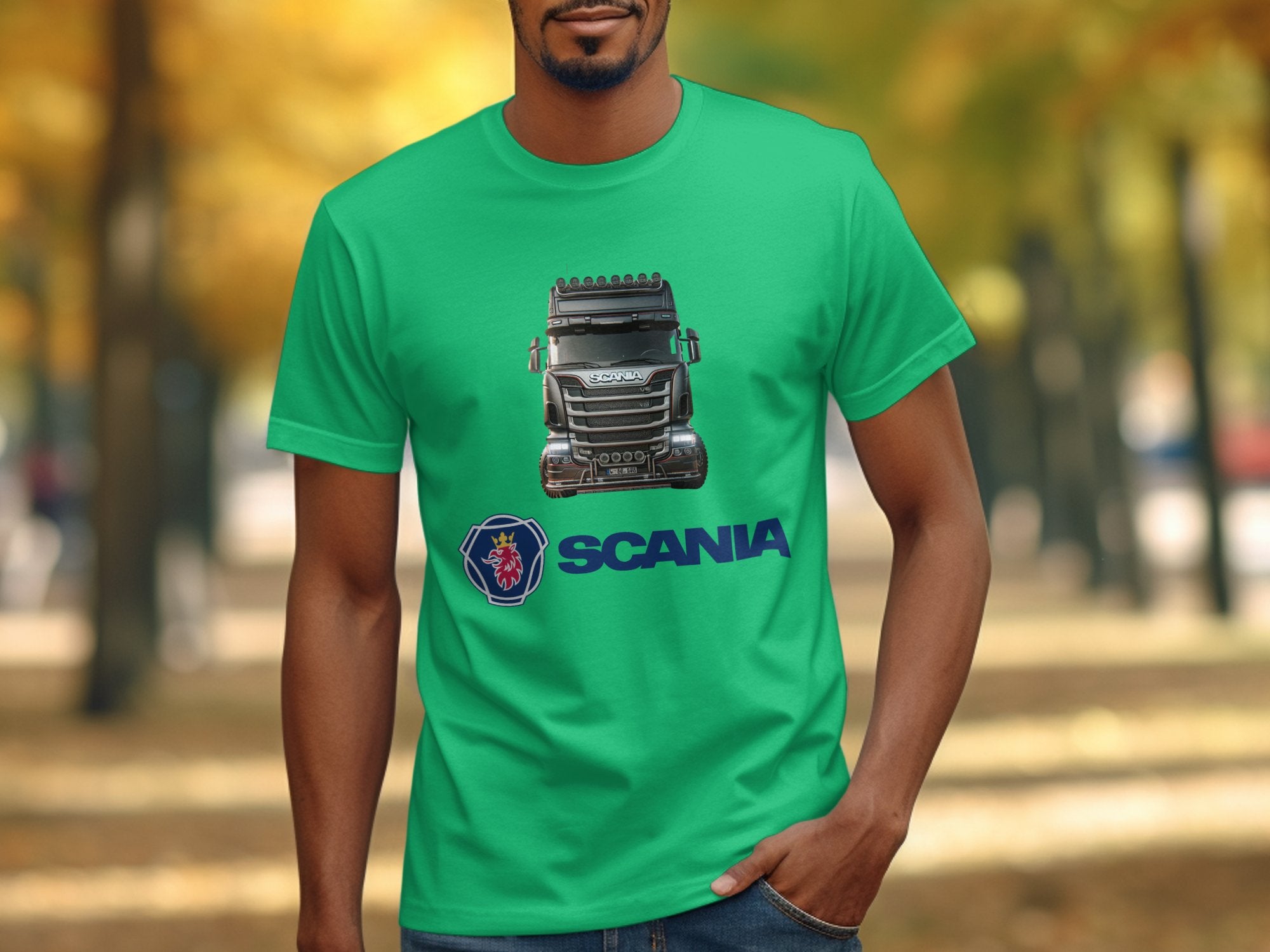 Someone wearing a bright green Garment Graphics T-shirt displaying a vibrant illustration of a Scania truck on the chest. SCANIA is boldly printed in blue below, with a tree-lined path and autumn leaves as the backdrop, ideal for fans of Scania trucks.