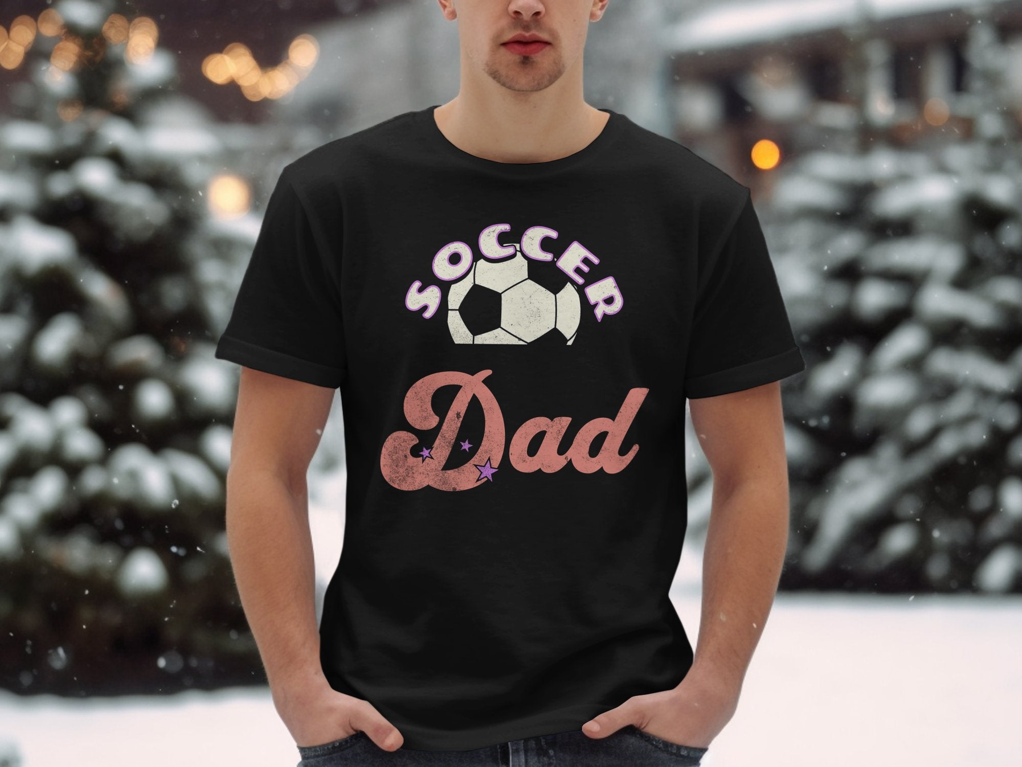 In a snowy outdoor setting, a person wears a Garment Graphics black graphic T-shirt with Soccer Dad and a soccer ball design. Snow-covered trees and softly lit lights complete the charming winter scene.