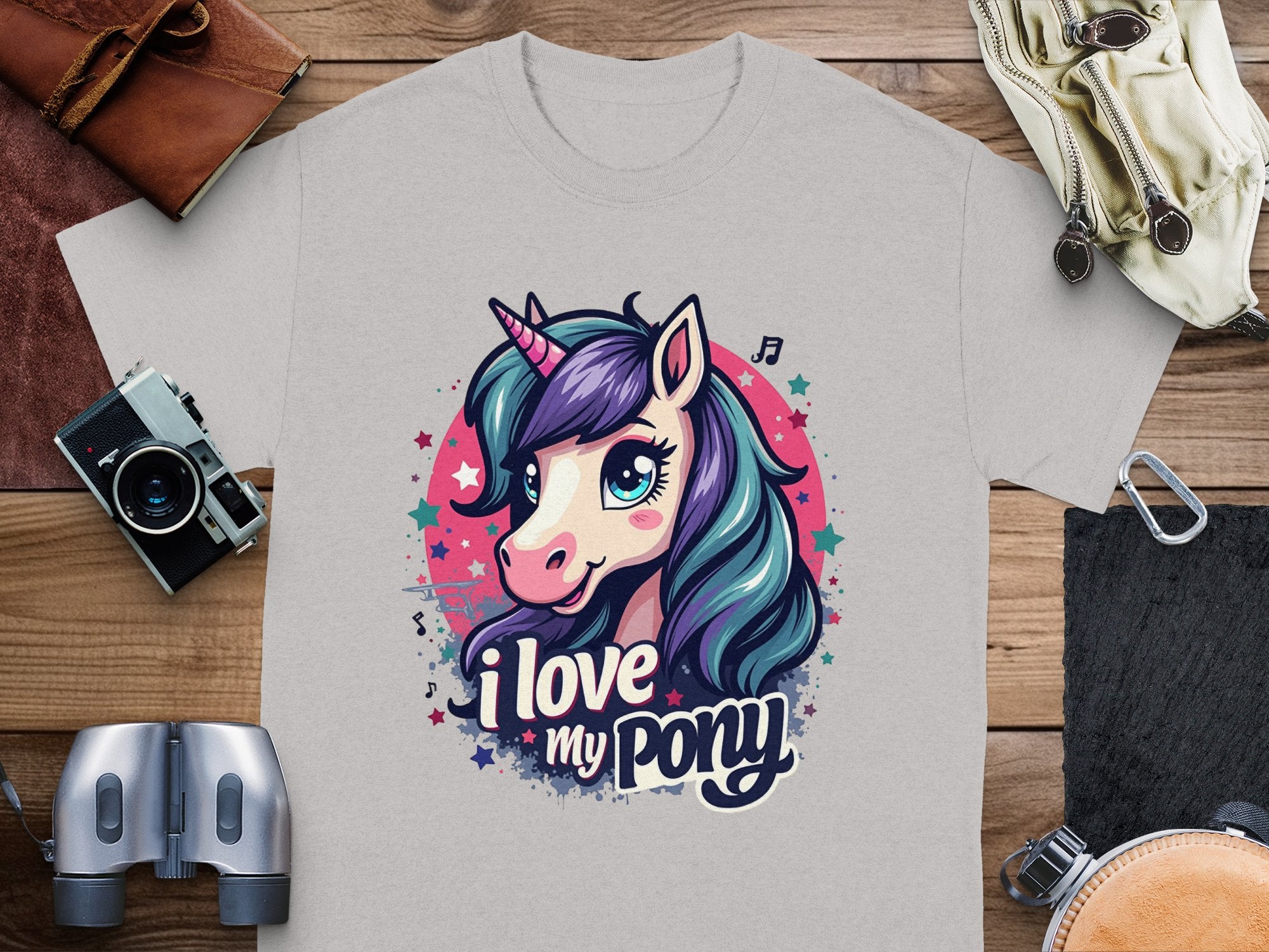 A gray Pony graphic T-shirt by Garment Graphics features a vibrant unicorn with purple and teal hair, displaying the phrase I love my pony. It’s perfect for unicorn enthusiasts and is pictured with a camera, binoculars, and leather pouch on a wooden surface.