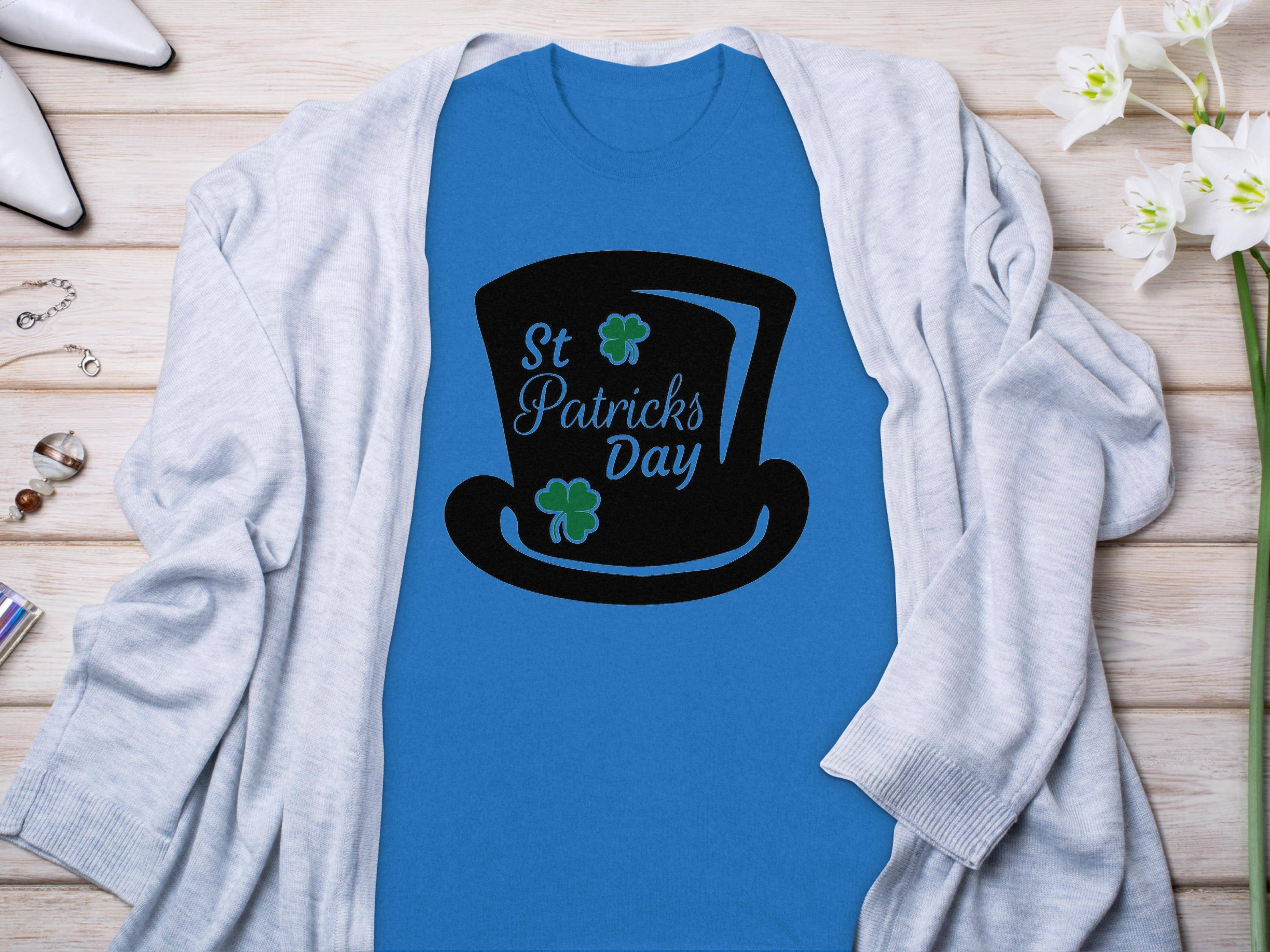 A Garment Graphics blue T-shirt featuring a black top hat, four-leaf clovers, and St Patricks Day sits on a wooden surface. Its surrounded by a light gray cardigan with white flowers and jewelry nearby.