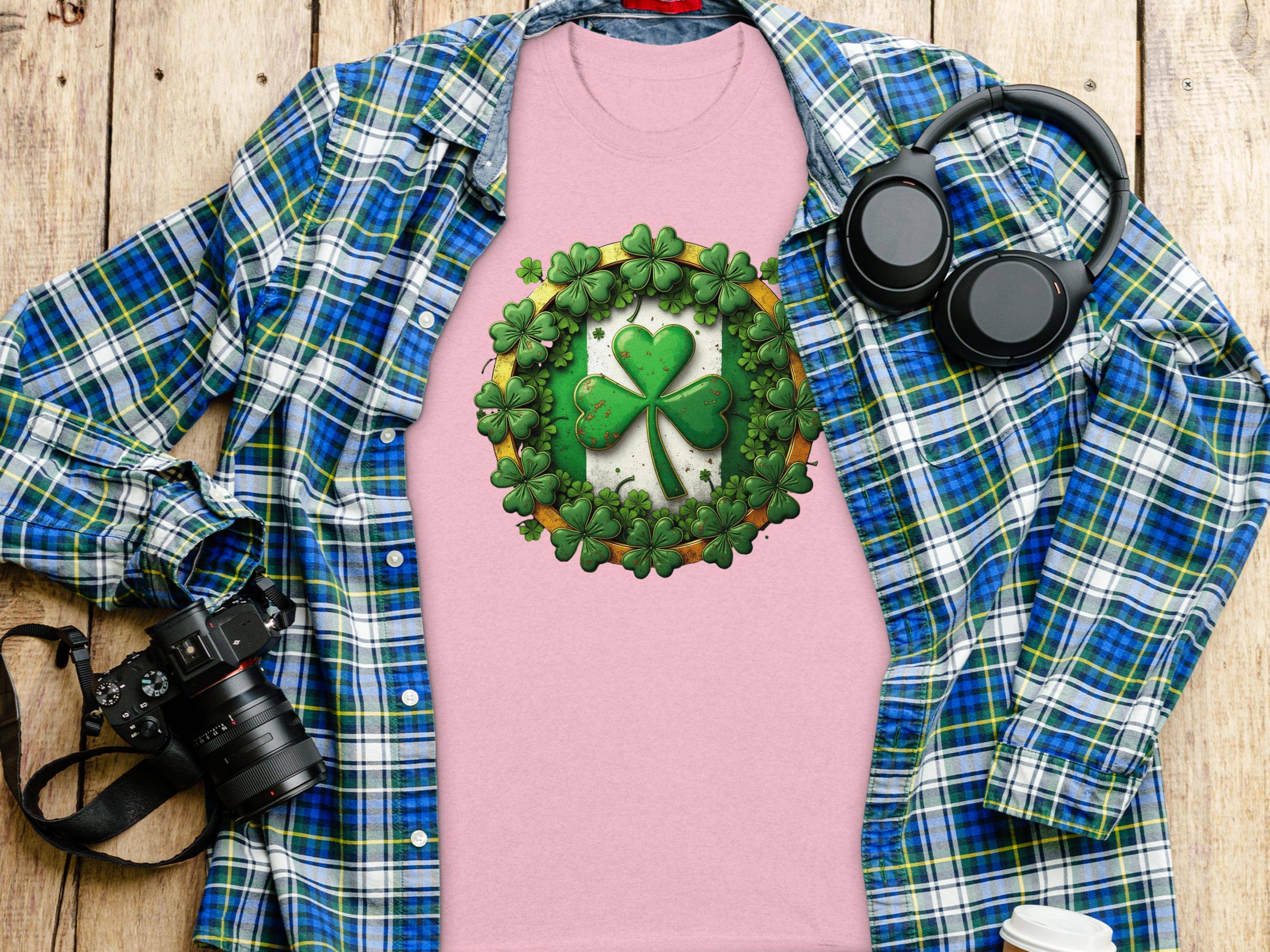 A Garment Graphics pink graphic T-shirt with a shamrock clover design sits on a wooden surface. Surrounding it are a blue plaid shirt, black headphones, a camera, and a white coffee cup lid—ideal for embracing the St. Patricks Day vibe in style.