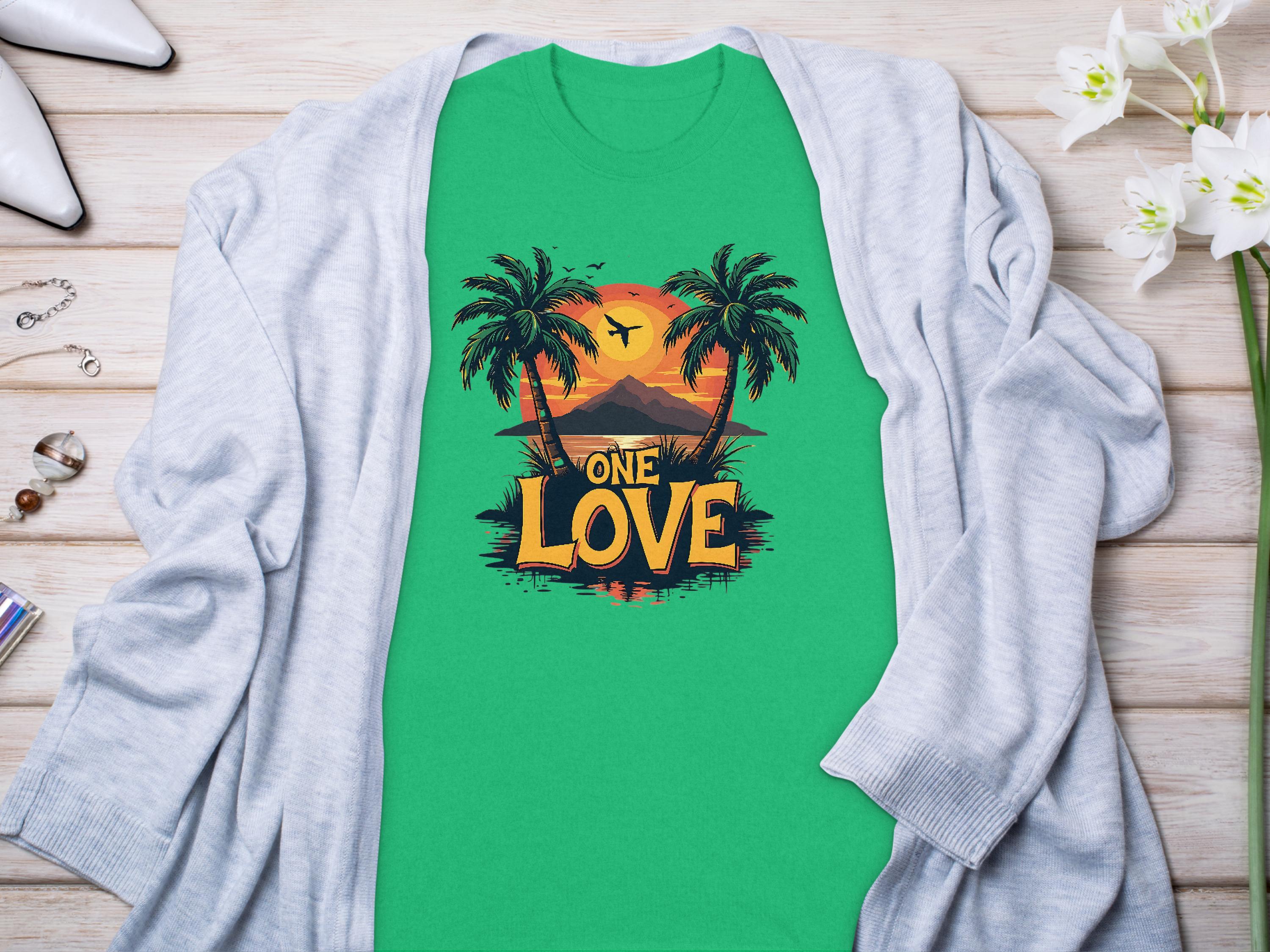 A Garment Graphics green T-shirt showcases a graphic with palm trees, a tropical sunset, and the text One Love. Styled with a light gray cardigan, its set beside white flowers and jewelry on wood—ideal for beach lovers.