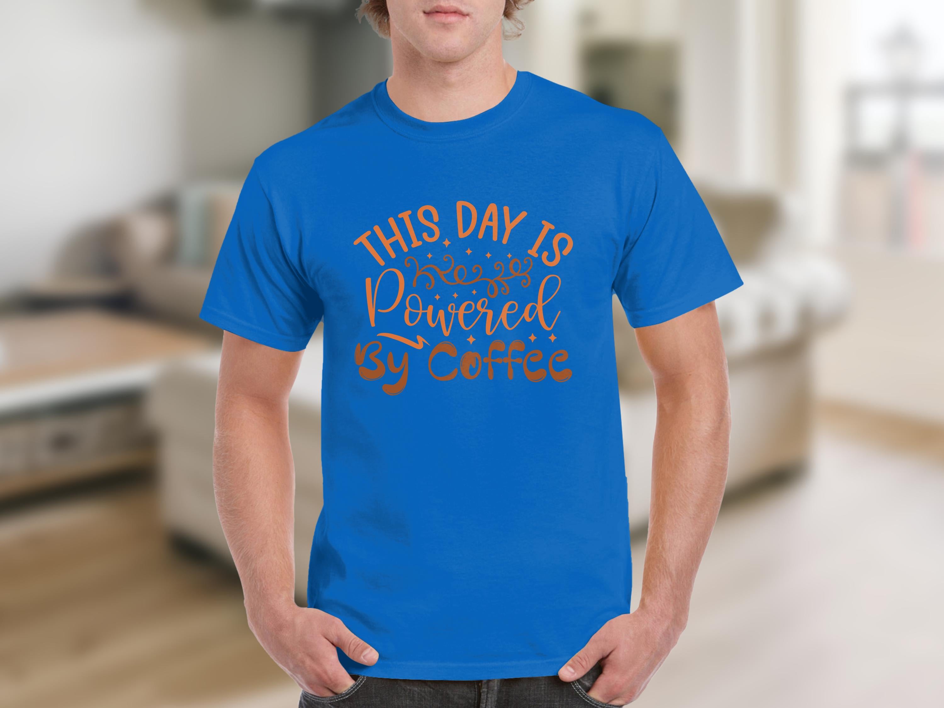 Indoors, a man wears a classic fit blue T-shirt by Garment Graphics, with This Day Is Powered by Coffee in decorative fonts. The blurred background of furniture like a sofa and shelves complements his positive, caffeine-inspired energy.