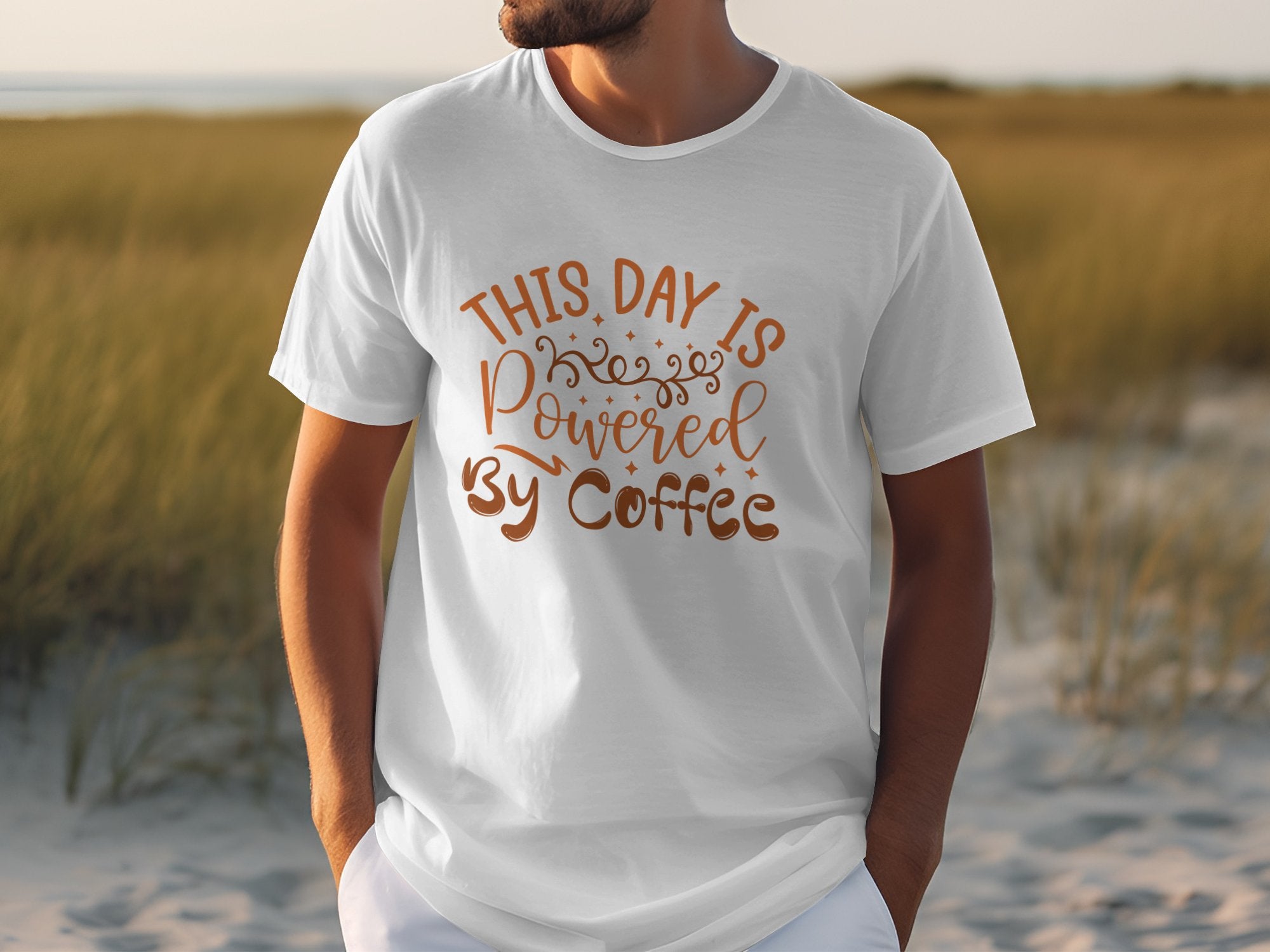 A man on the beach wears a classic fit white t-shirt by Garment Graphics with This day is powered by coffee in bold brown letters, radiating positivity against a serene coastal backdrop of soft sand and tall grass.