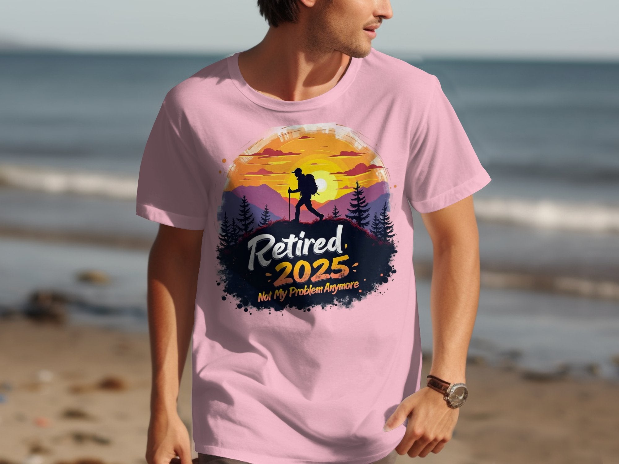 On a beach, a man wears a Garment Graphics T-shirt with a pink sunset displaying Retired 2025: Not My Problem Anymore and a hiker silhouette. The gentle ocean waves symbolize his peaceful embrace of retirement.