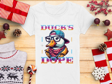 A white T-shirt by Garment Graphics showcases a cartoon duck with swag, wearing glasses, a cap, and a leather jacket. Ducks Dope is in colorful letters. Its displayed on wood with holiday decorations and wrapped gifts.