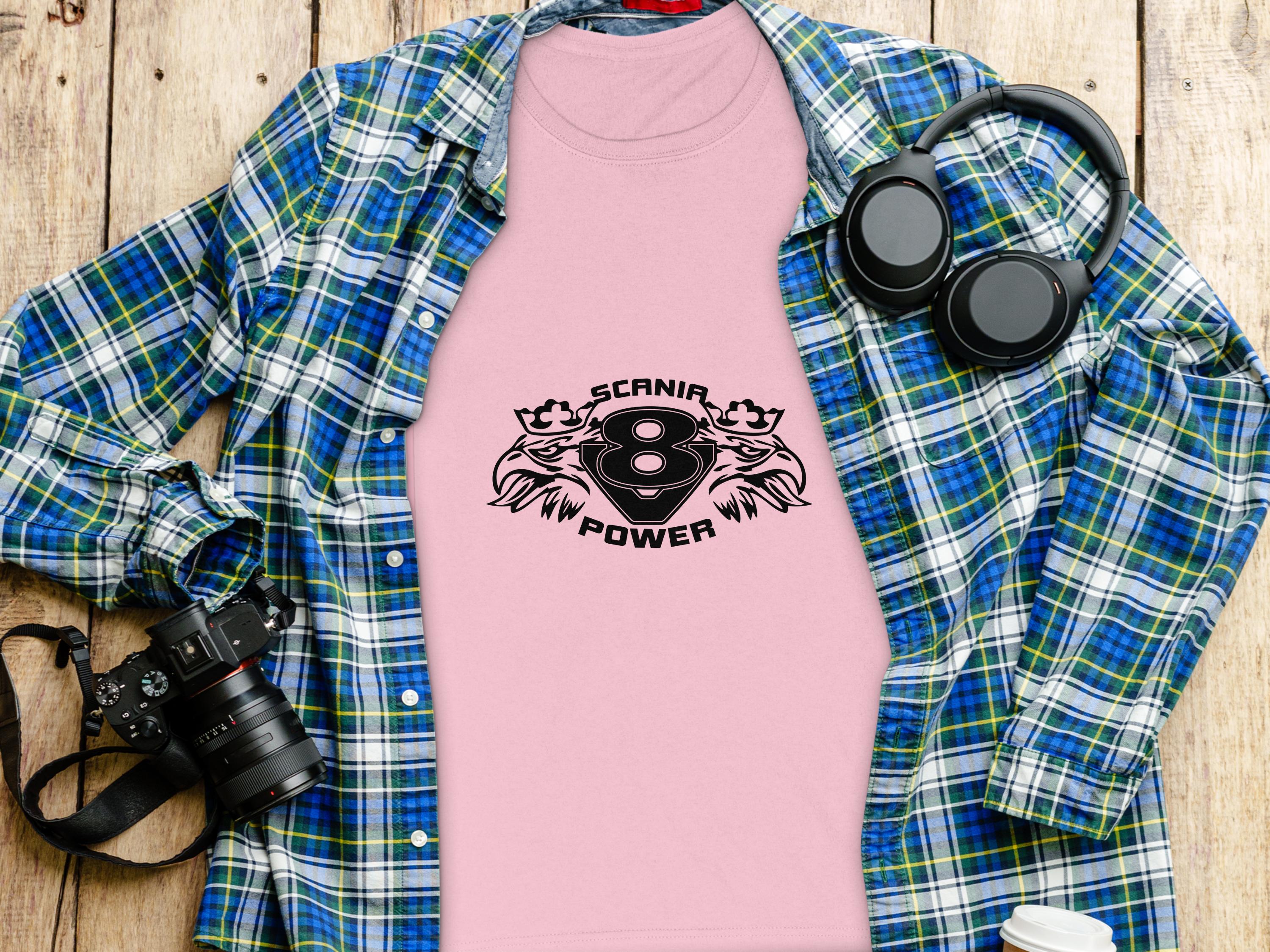 A pink Garment Graphics t-shirt with Scania V8 Power and an eagle head design sits on a wooden surface, paired with a blue plaid shirt, black headphones, camera, and a white coffee cup lid.