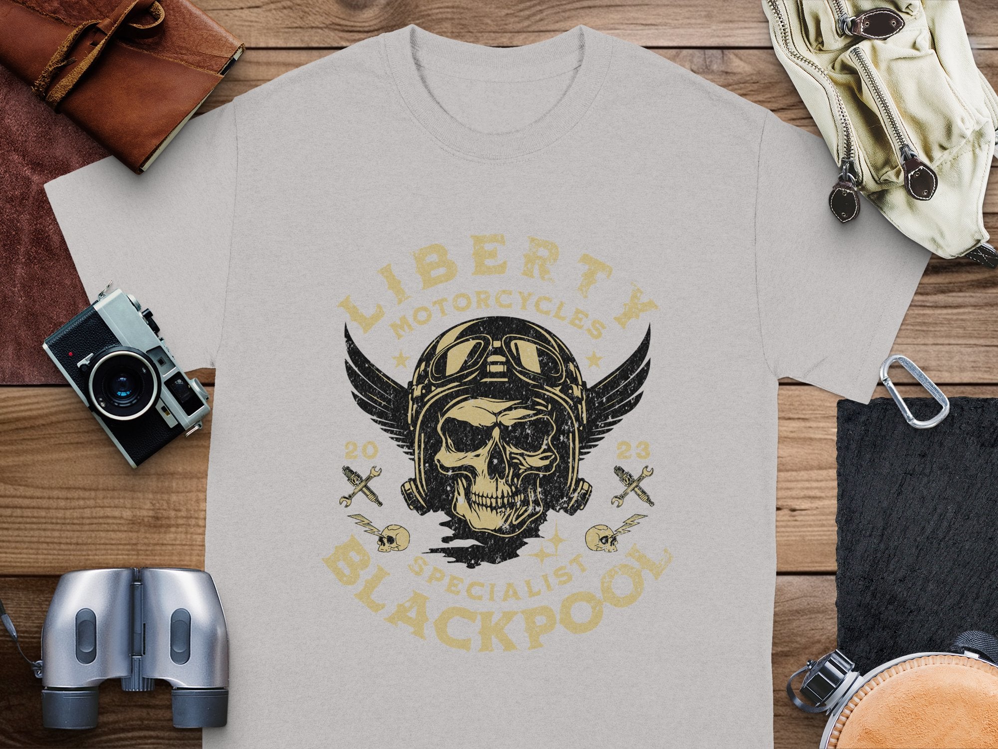A grey Garment Graphics T-shirt lies on a wooden surface featuring a vintage design: a skull in aviator goggles and helmet with Liberty Motorcycles Specialist Blackpool 2023 text. Ideal for motorcycle enthusiasts, its paired with a camera, binoculars, leather wallet, gloves, and beige cap.