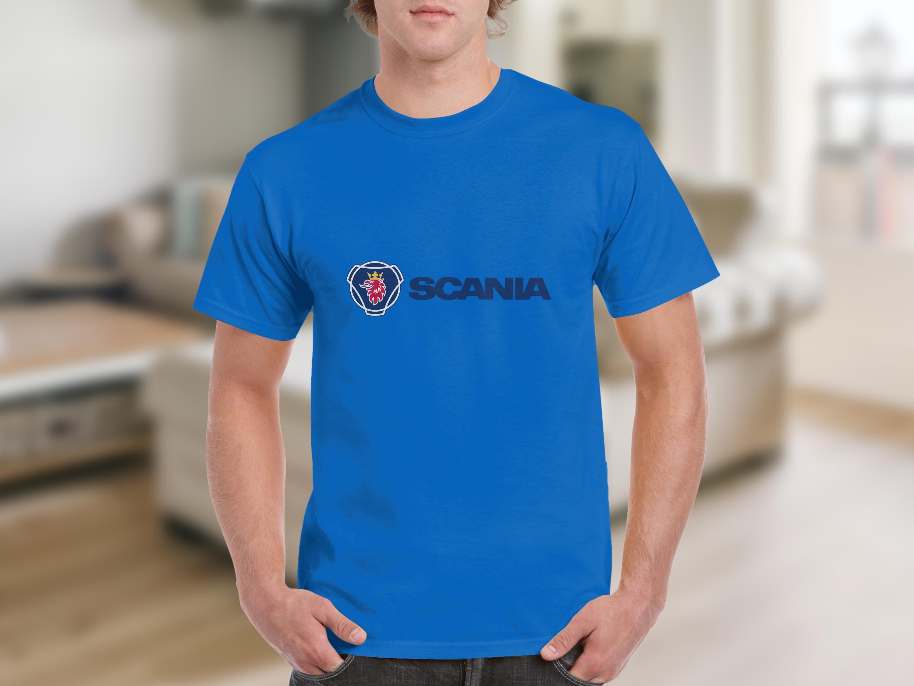 Indoors, a person sports a blue cotton T-shirt by Garment Graphics with the Scania logo prominently displayed. The blurred background features a cozy sofa and furniture, capturing the essence of incorporating Scania merchandise into everyday life.