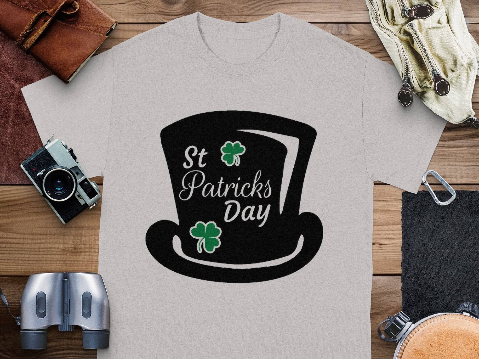 Garment Graphics grey T-shirt features a black top hat, St Patricks Day text, two green shamrocks, and a four-leaf clover theme surrounded by a camera, binoculars, leather bags, and more on wood.