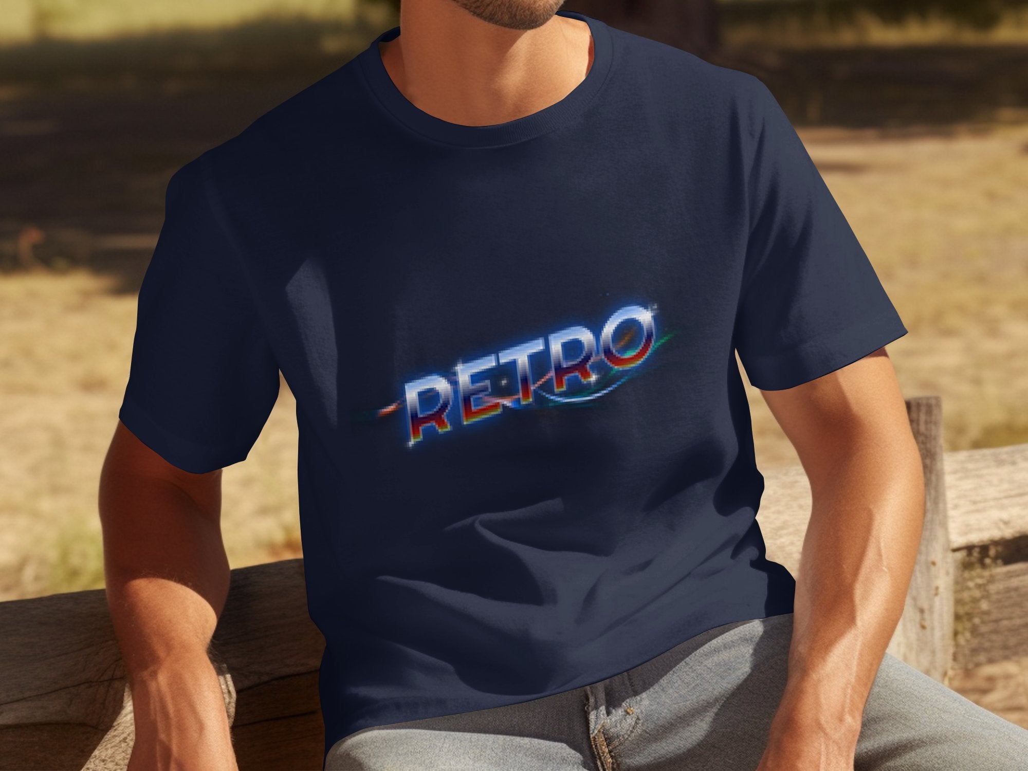 A person sits on a wooden bench wearing a Garment Graphics navy blue t-shirt with RETRO in colorful, stylized letters across the chest. The shirt captures vintage charm against a blurred natural backdrop, though the face is not visible.