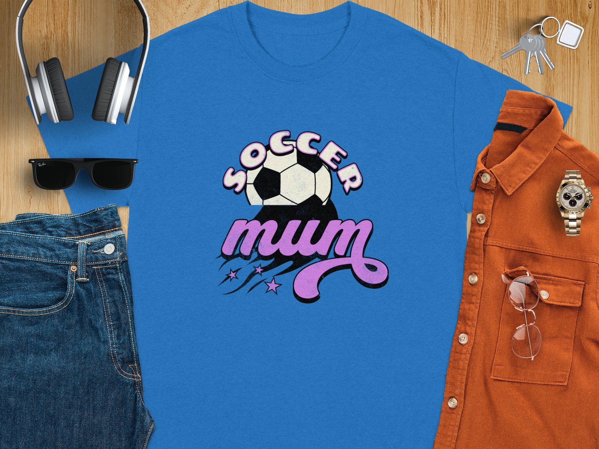 The classic fit blue t-shirt from Garment Graphics features Soccer Mum in pink and white, with a soccer ball graphic. Displayed on wood, its styled with headphones, sunglasses, jeans, an orange button-up shirt, a watch, and keys.