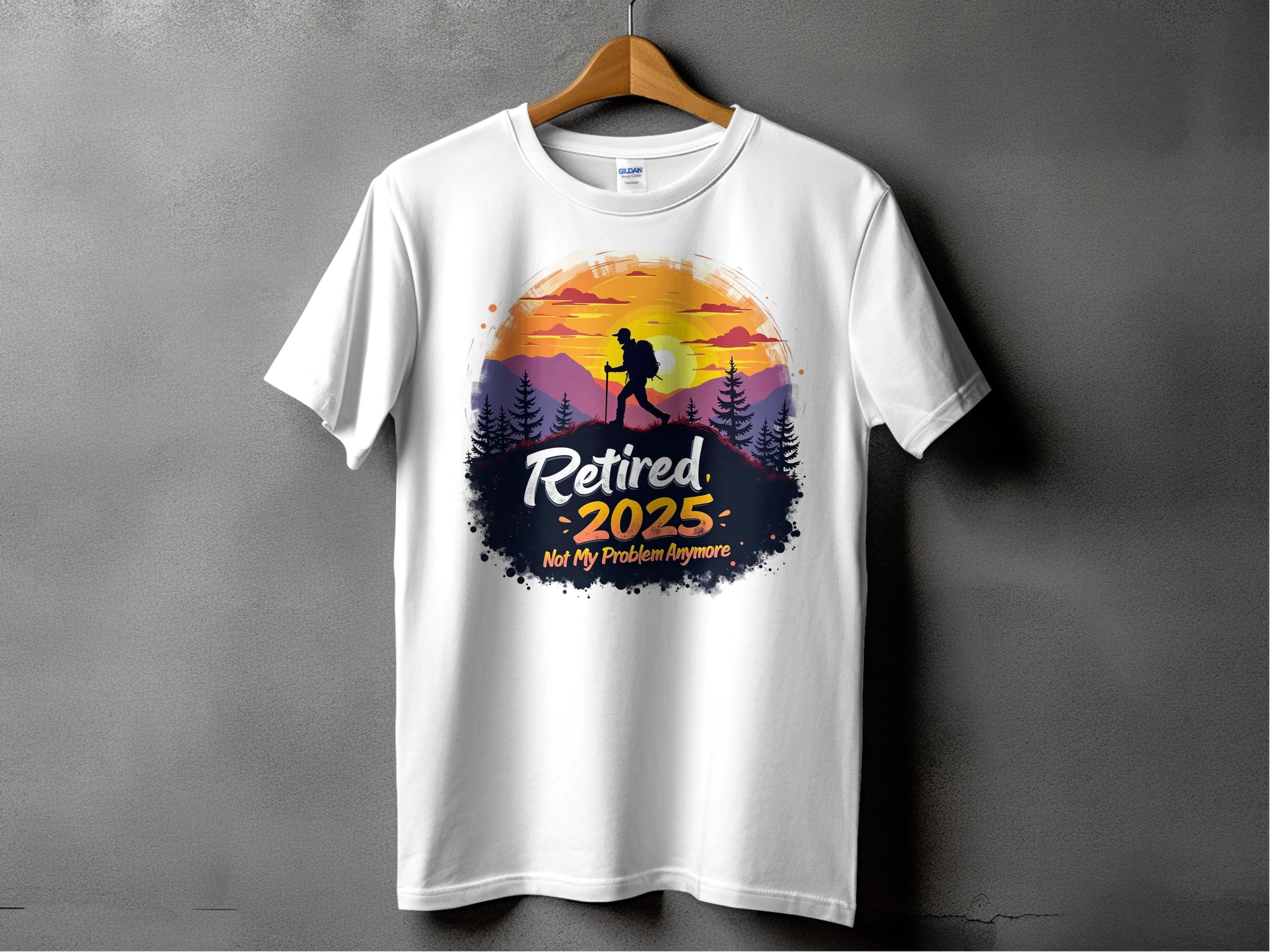 A white Garment Graphics t-shirt hangs on a wooden hanger, featuring a vibrant sunset with a hiker silhouette and bold text: Retired 2025 - Not My Problem Anymore, embodying the spirit of carefree retirement.