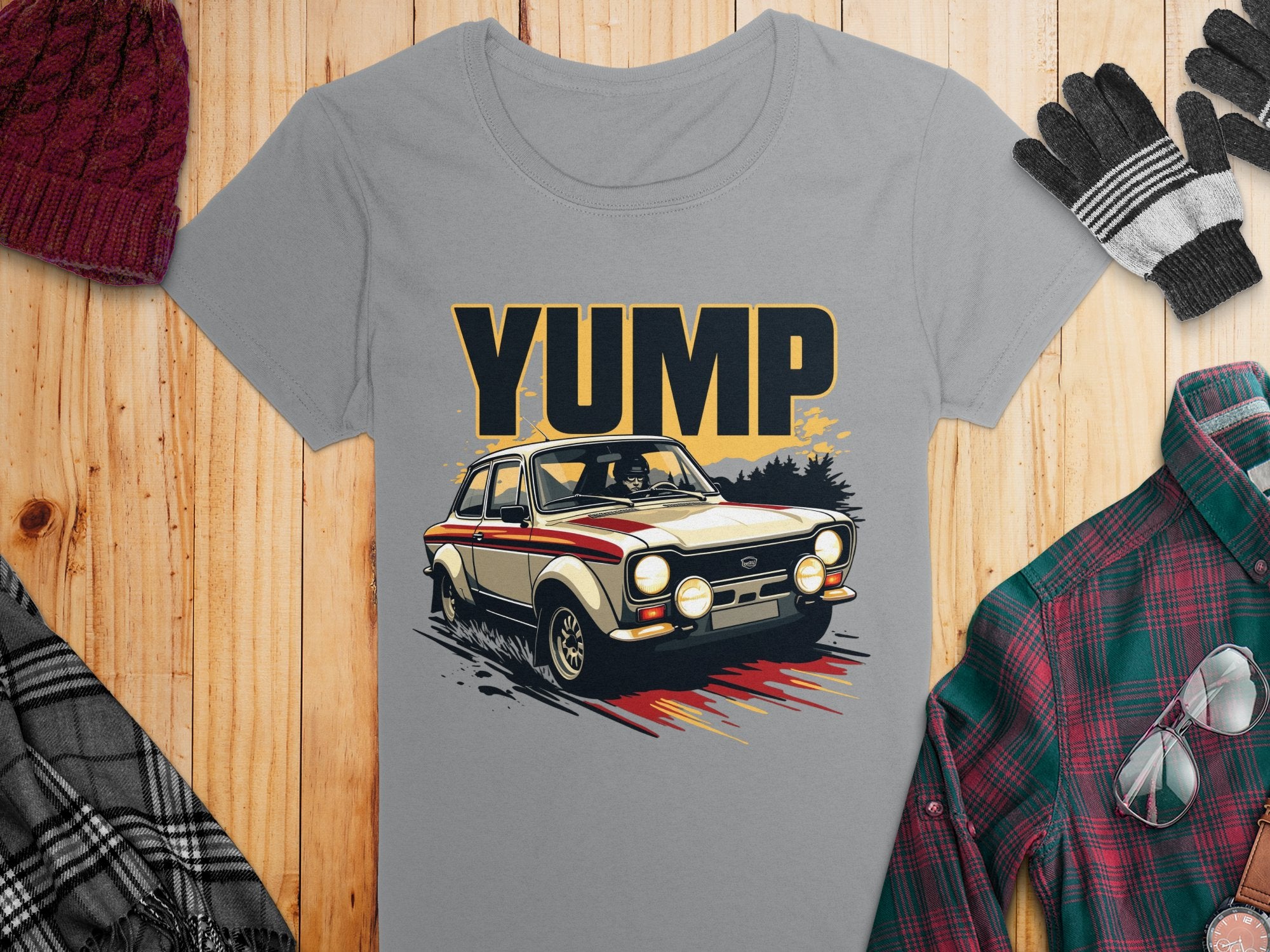 A gray Garment Graphics T-shirt with a YUMP vintage car design lays on wood, surrounded by a red knit hat, gloves, plaid scarf, and green/red plaid shirt—a retro T-shirt lovers dream.