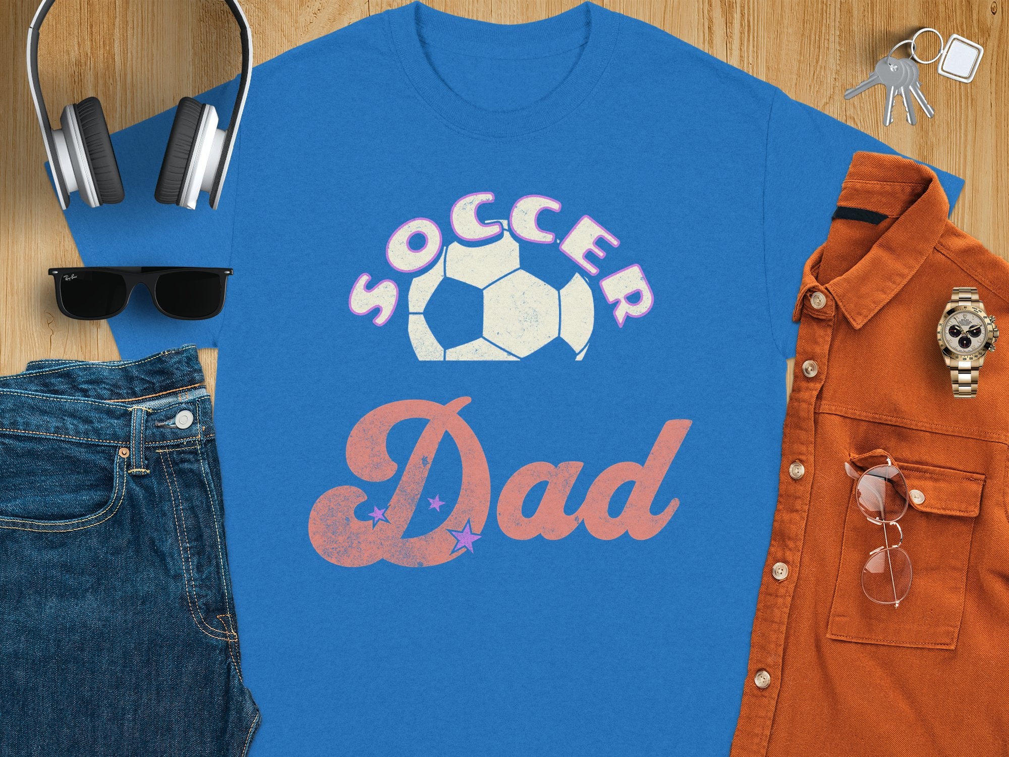 A Garment Graphics blue graphic T-shirt with SOCCER Dad and a soccer ball design is on a wooden surface, surrounded by sunglasses, denim jeans, headphones, a brown button-up shirt, a watch, wallet, and keys.