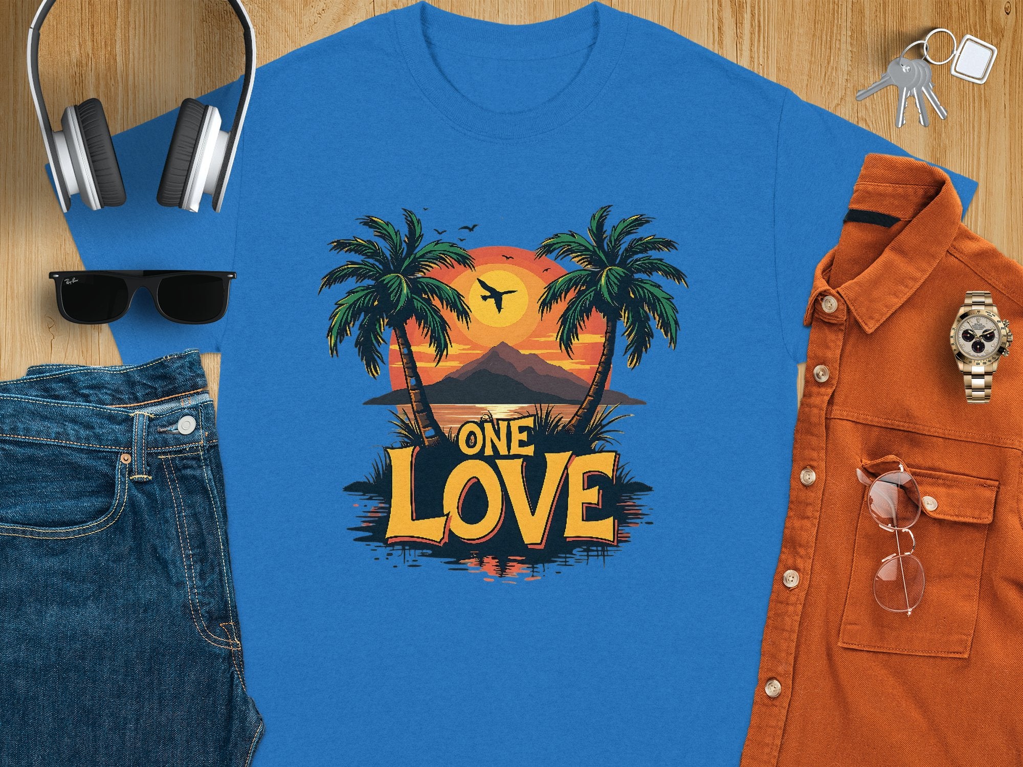 A flat lay showcases a blue Garment Graphics T-shirt with a One Love tropical sunset print. It includes palm trees and a sunset, ideal for beach lovers. Surrounding items are denim jeans, sunglasses, a brown shirt, wristwatch, headphones, wallet, and keys on wood.