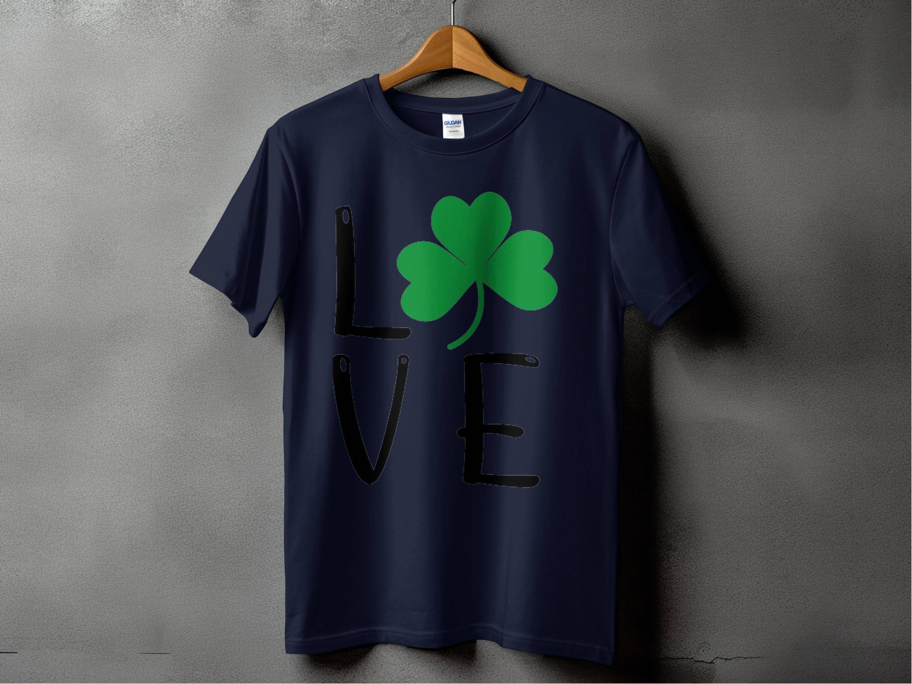 A Garment Graphics T-shirt, dark blue and hanging on a wooden hanger against a gray background, embodies Irish spirit. The word LOVE features a clover graphic as the O, making it ideal for a St. Patricks Day collection.