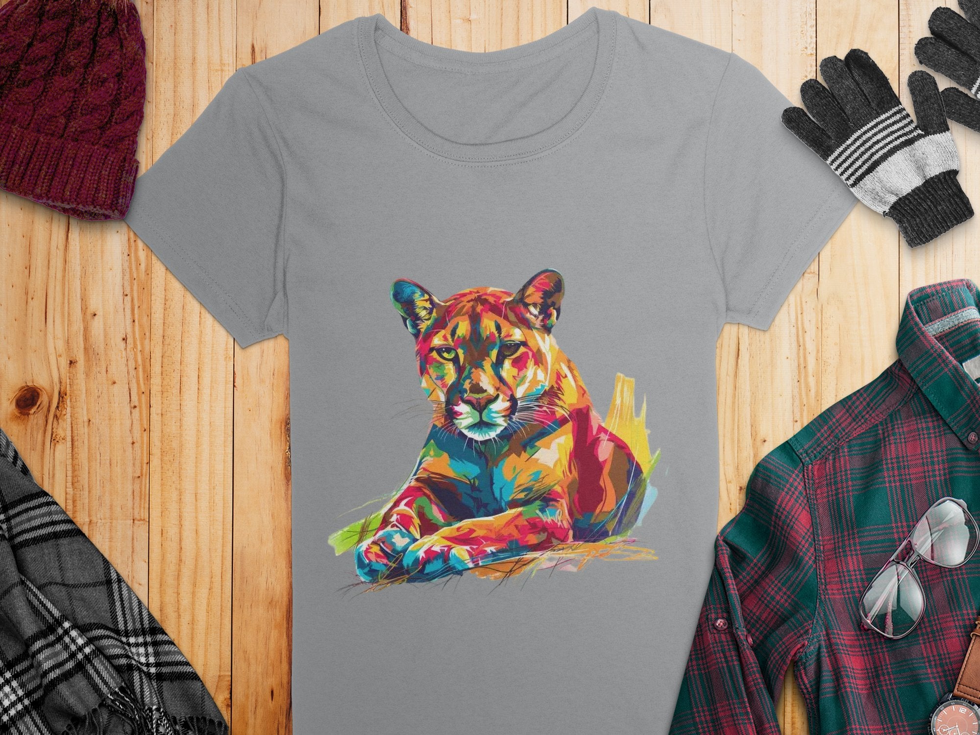 A Garment Graphics gray T-shirt featuring a colorful puma design rests on a wooden surface, surrounded by a maroon knit beanie, black and gray gloves, a plaid scarf, and a green and red flannel shirt.