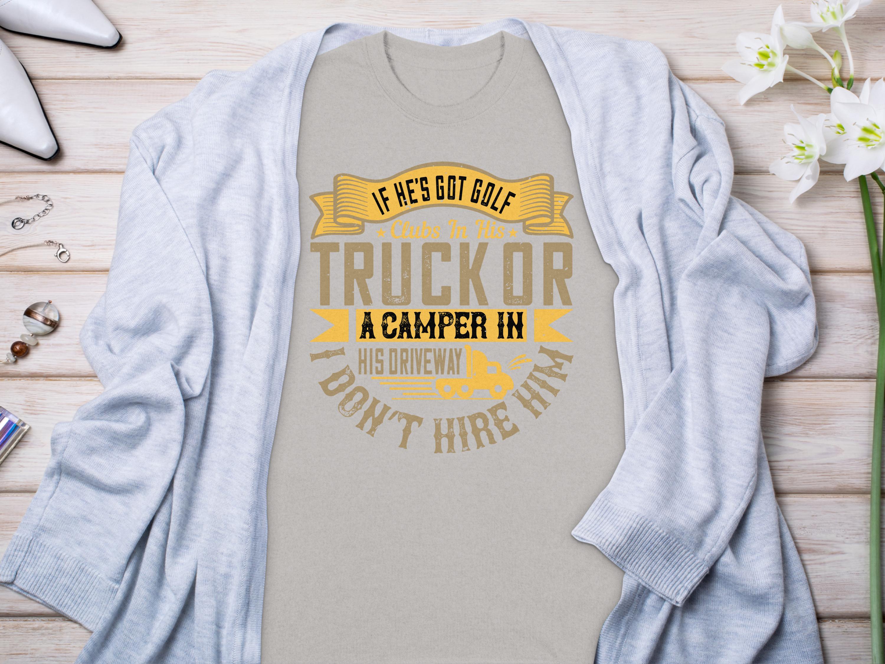 Displayed on a wooden surface, Garment Graphics gray t-shirt features the humorous quote, If hes got golf clubs in his truck or a camper in his driveway, I dont hire him, surrounded by a light cardigan, flowers, and accessories—an ideal gift for humor enthusiasts.