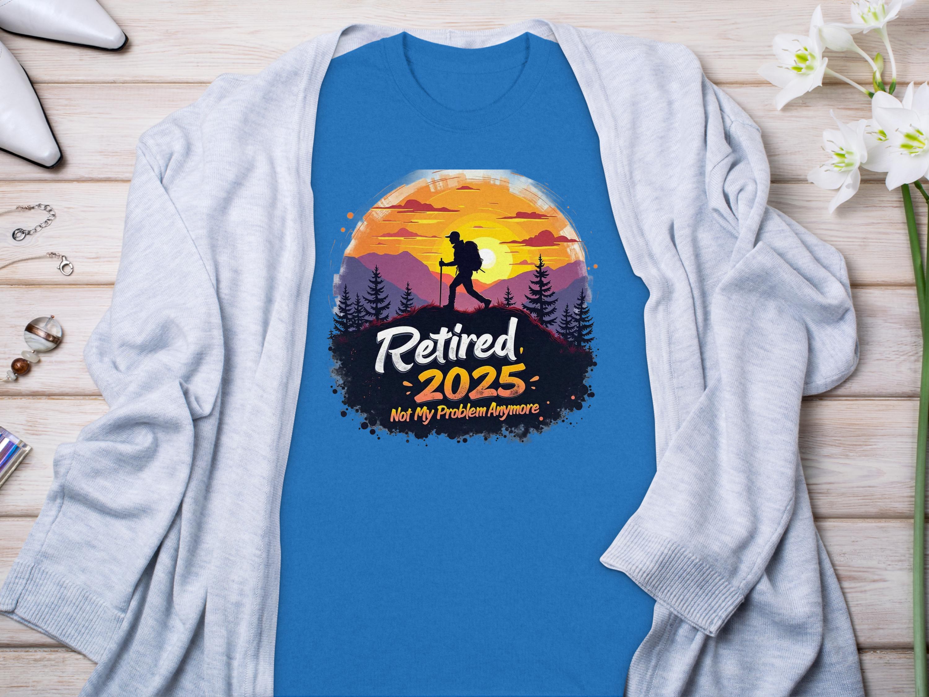 The Garment Graphics blue Retired 2025, Not My Problem Anymore sunset t-shirt with a hiker silhouette pairs perfectly with a light gray cardigan. Surrounded by elegant accessories and white flowers, its ideal for embracing retirement in style.