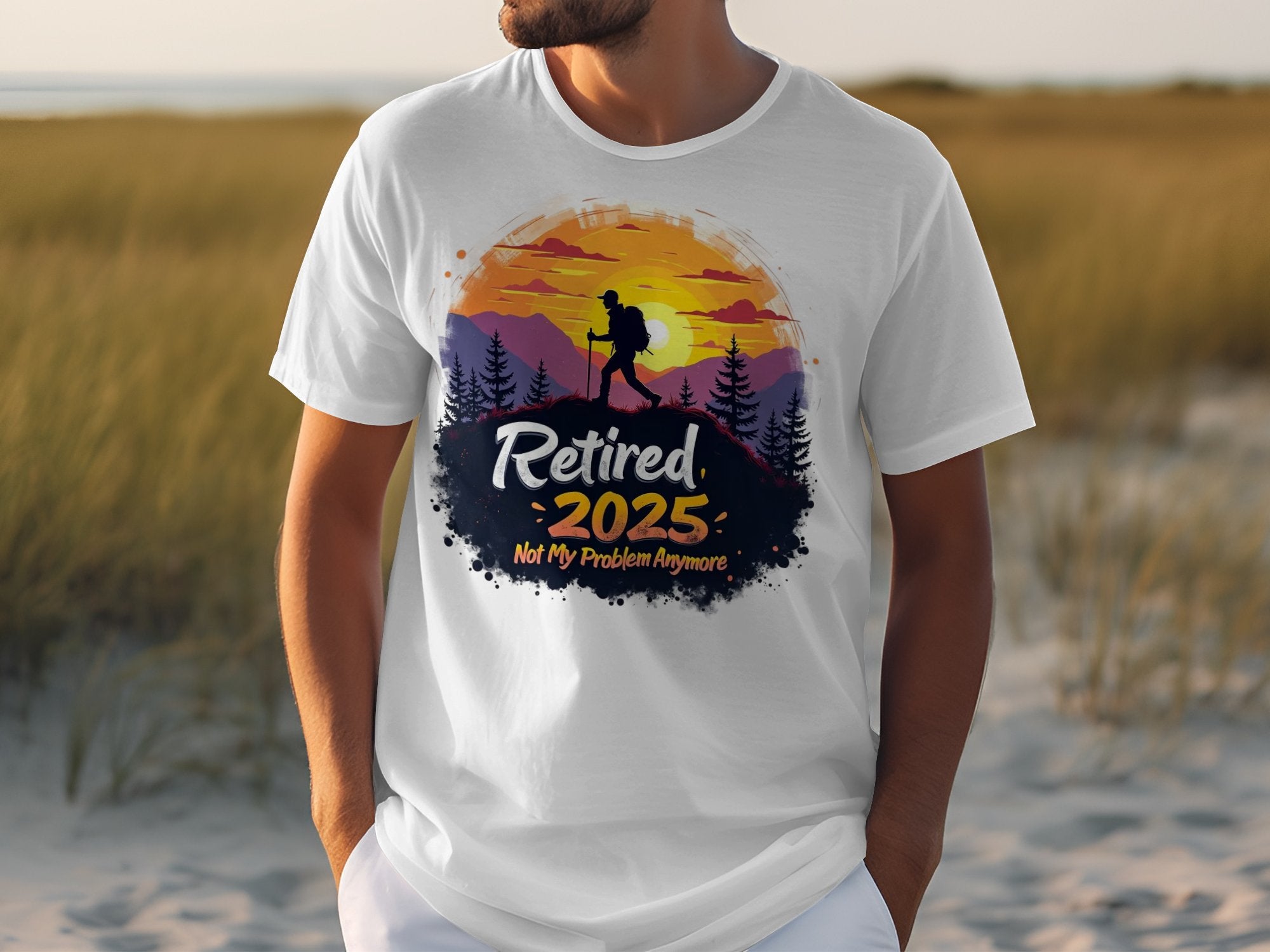 A person is wearing a Garment Graphics white T-shirt featuring a colorful design of a hiker silhouette with mountains and trees, alongside the text Retired 2025 Not My Problem Anymore, embodying retirement vibes. A grassy area sets the backdrop for this lively scene.