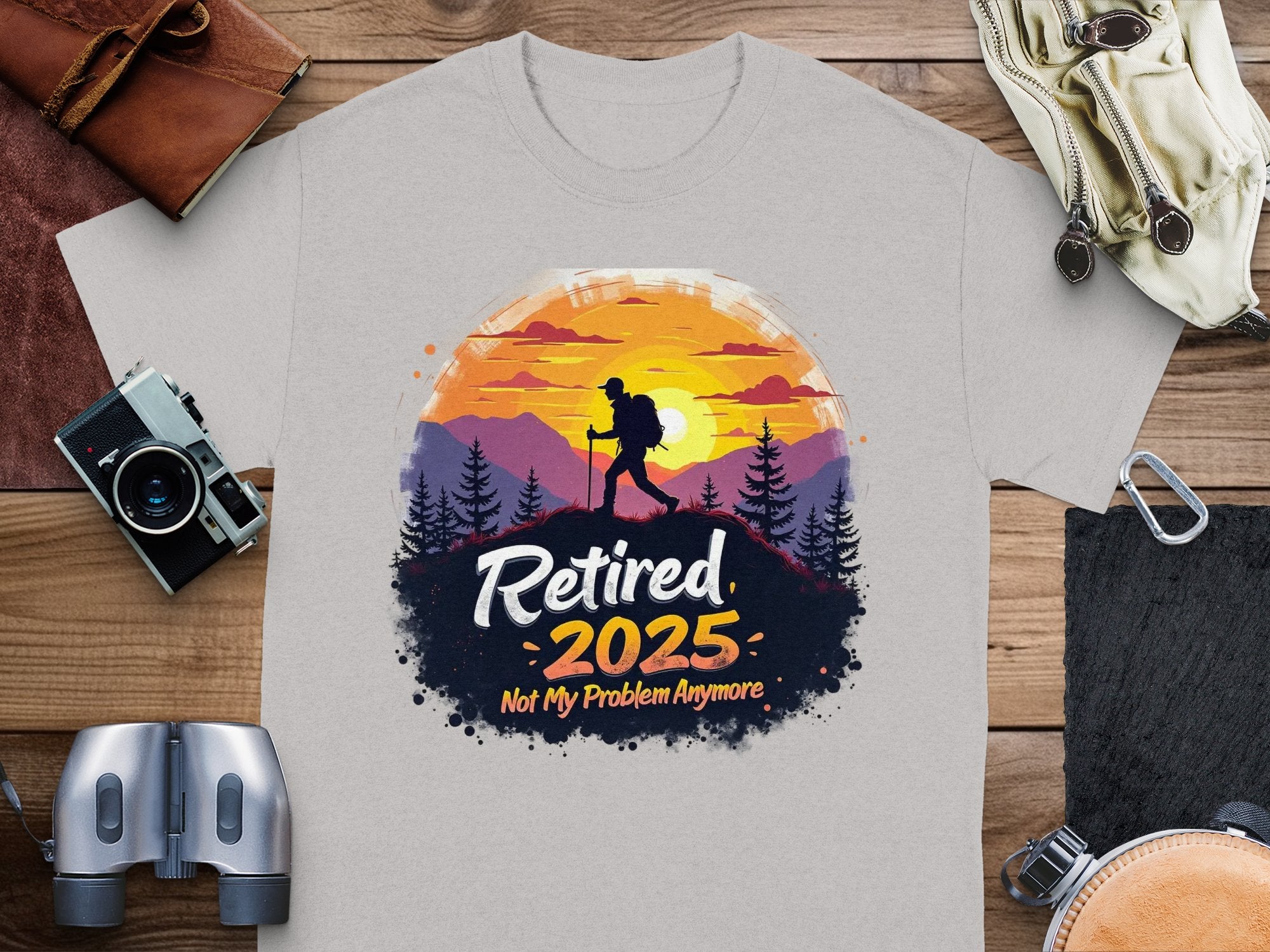 A gray T-shirt by Garment Graphics showcases a hiker with a vibrant sunset and tree frame, featuring Retired 2025: Not My Problem Anymore. Placed on wood alongside essentials like a camera and binoculars, its ideal for those nearing retirement.