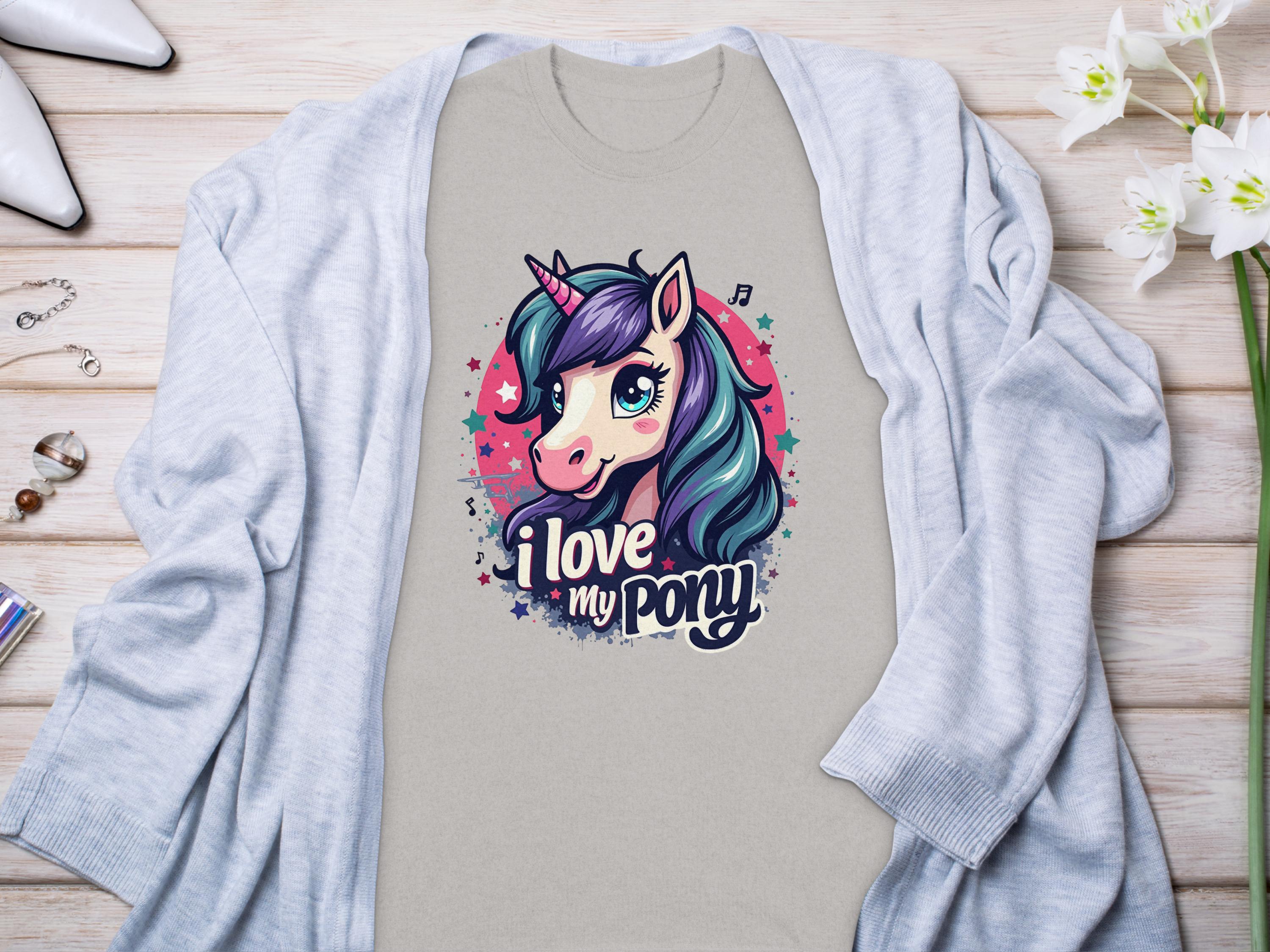 A beige pony graphic T-shirt by Garment Graphics features a vibrant unicorn with the phrase I love my pony. Paired under a light gray cardigan on light wood, its beautifully accented with flowers and small accessories, ideal for any unicorn enthusiast.