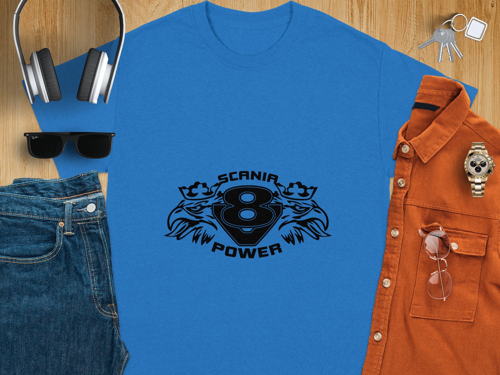 A Garment Graphics blue t-shirt with Scania 8 Power text and graphic design on a wooden surface, surrounded by wireless headphones, denim hat, sunglasses, blue jeans, orange shirt, wristwatch, and keys.