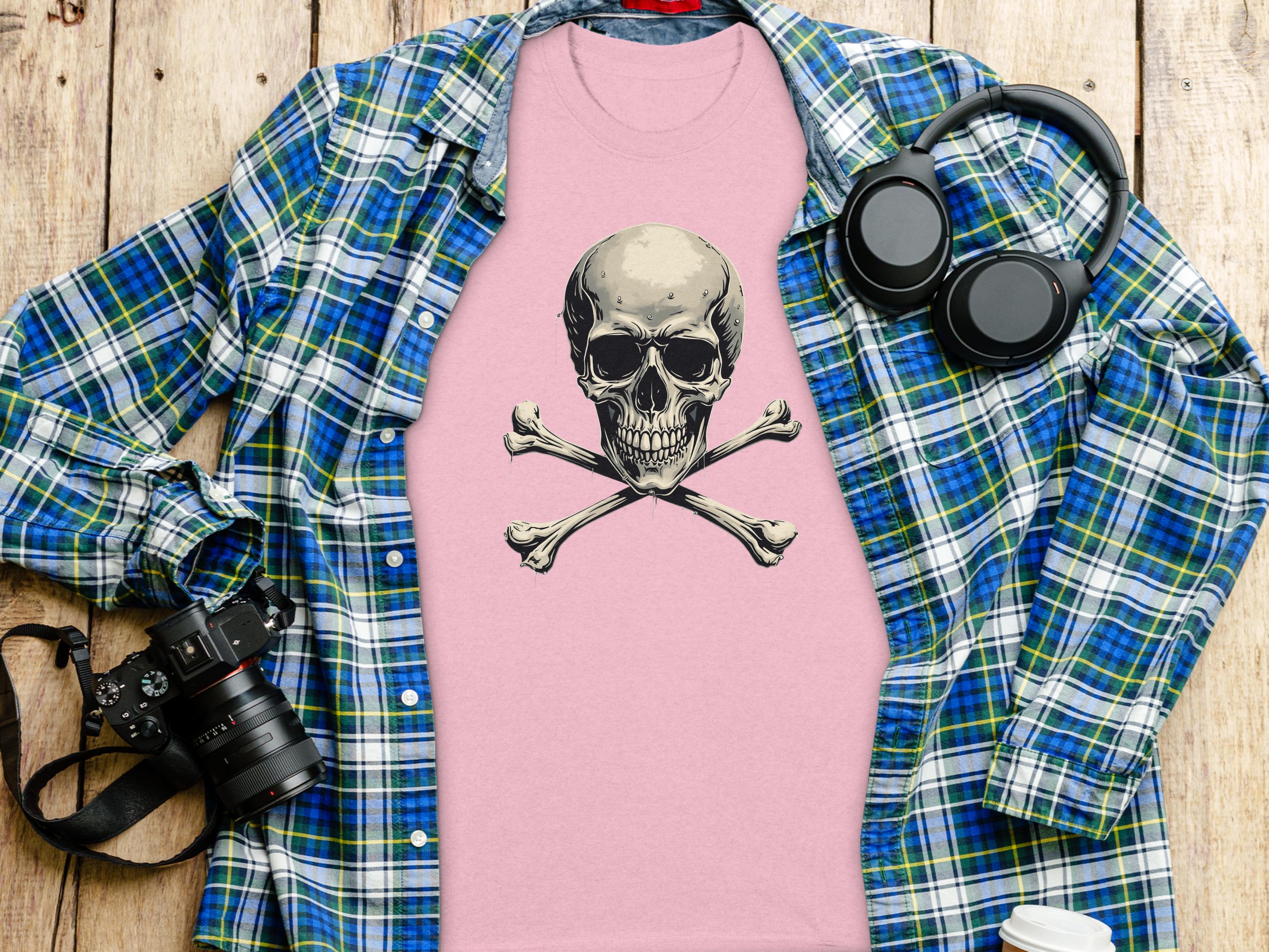 A flat lay image presents a striking look with Garment Graphics pink graphic t-shirt featuring a skull and crossbones. Its accompanied by a blue plaid shirt, headphones, a camera, and a takeaway coffee cup on a wooden surface.