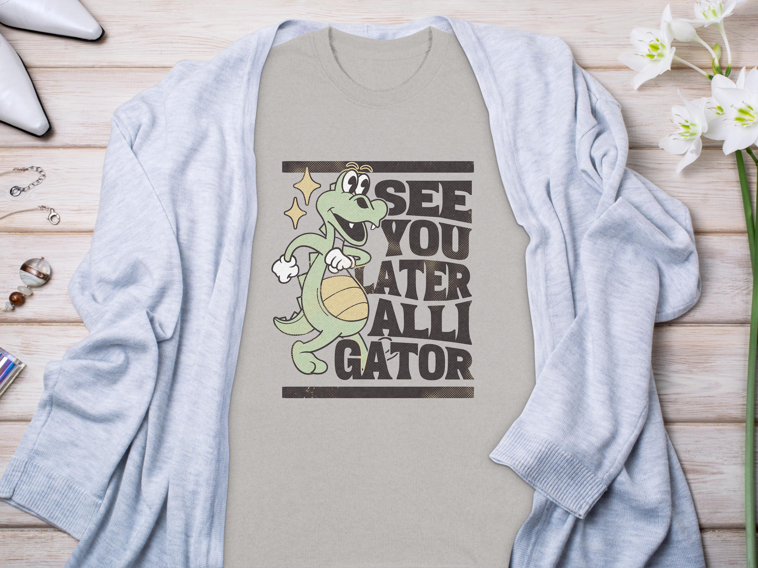 Garment Graphics presents a casual t-shirt featuring a cartoon alligator with a basketball and the phrase See You Later Alligator. Styled on wood, its paired with a light gray cardigan, flowers, and accessories for a relaxed vibe.