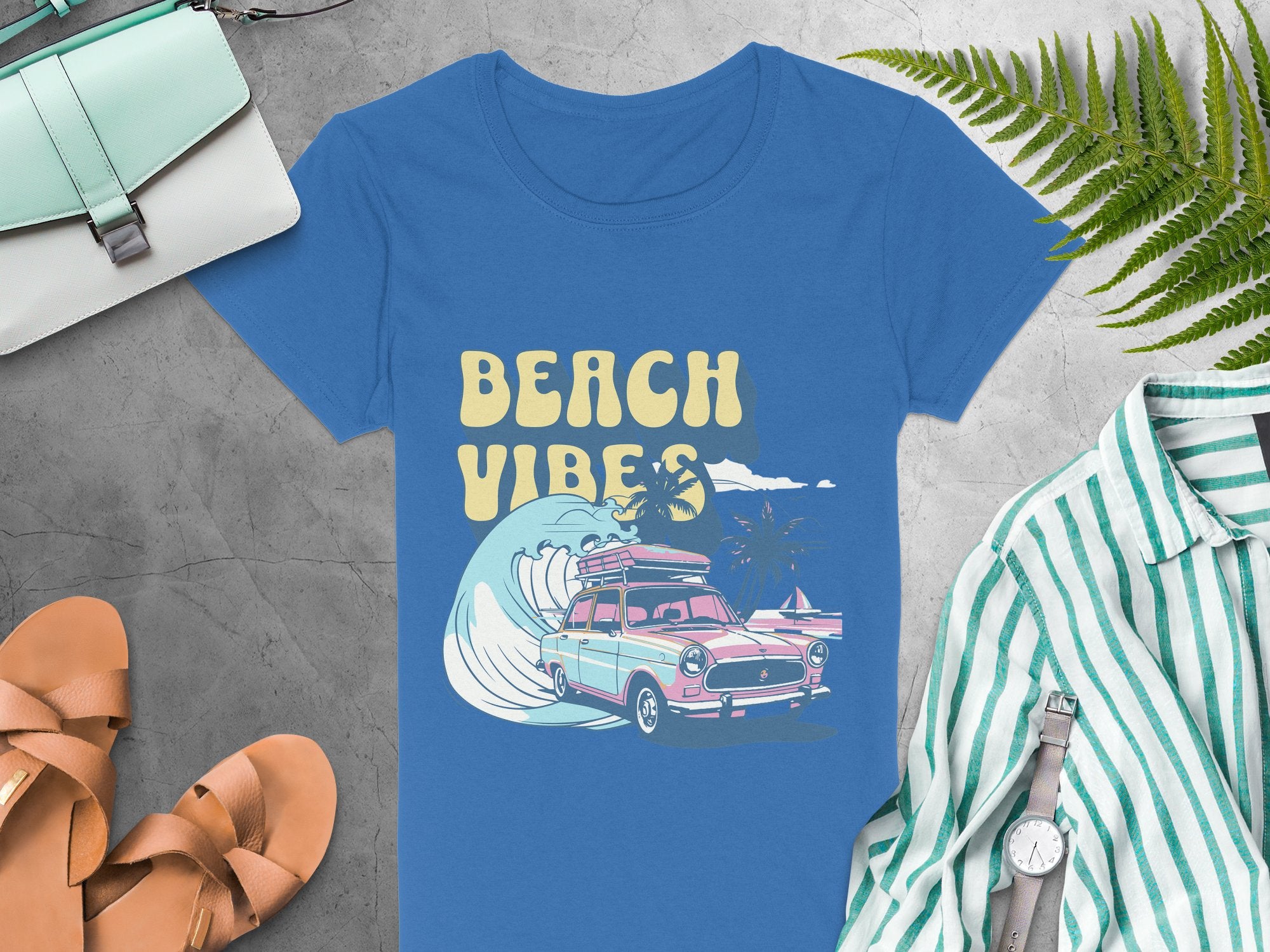A retro surf blue t-shirt by Garment Graphics displays a vintage-style graphic of a pink car with surfboards and BEACH VIBES text. It lies near tan sandals, a green and white striped shirt, watch, purse, and green fern leaves on gray surface.