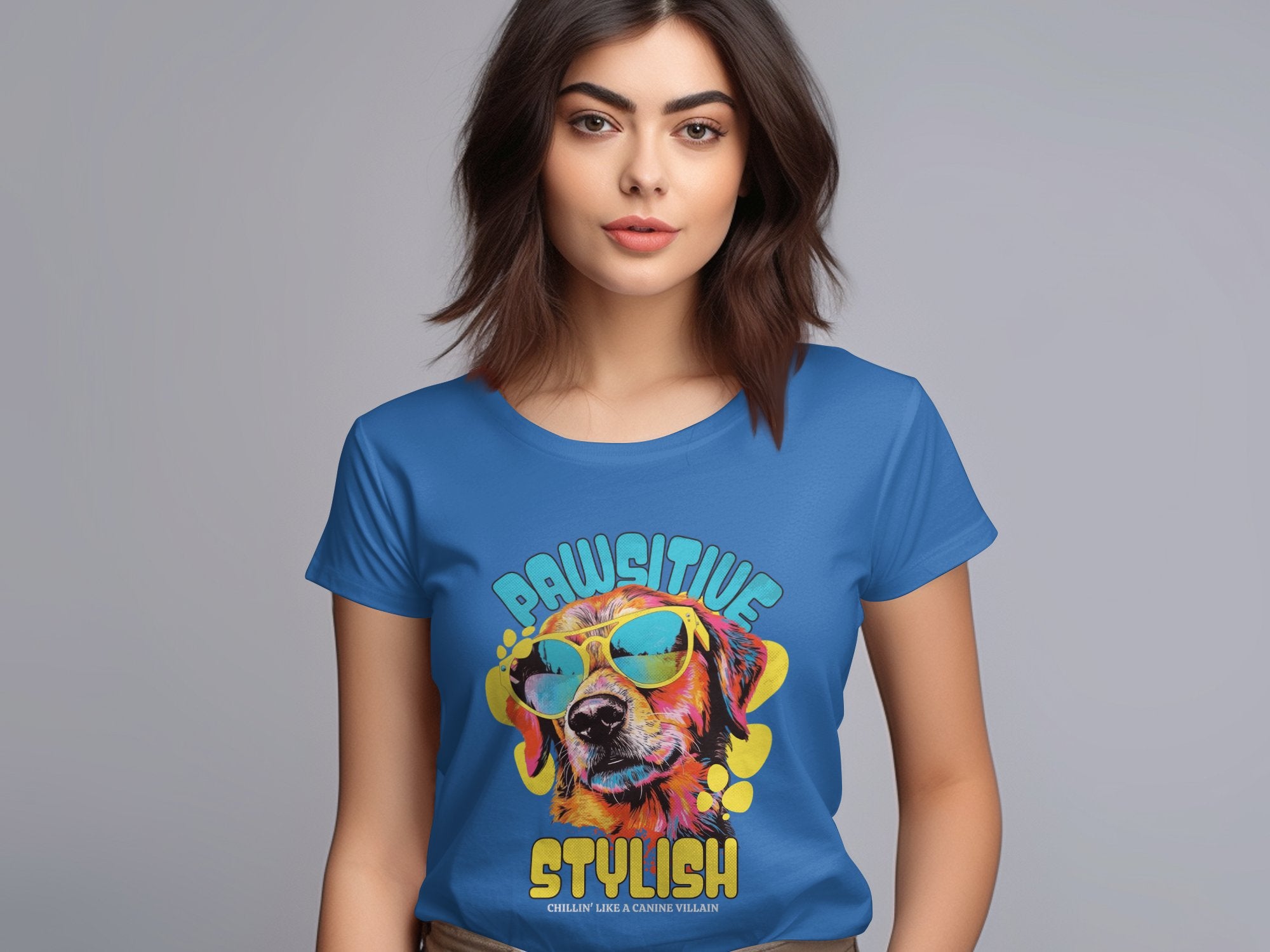 A woman with shoulder-length brown hair wears a Garment Graphics womens blue t-shirt featuring a graphic of a dog in sunglasses and the words Pawsitive Stylish on a plain gray background.