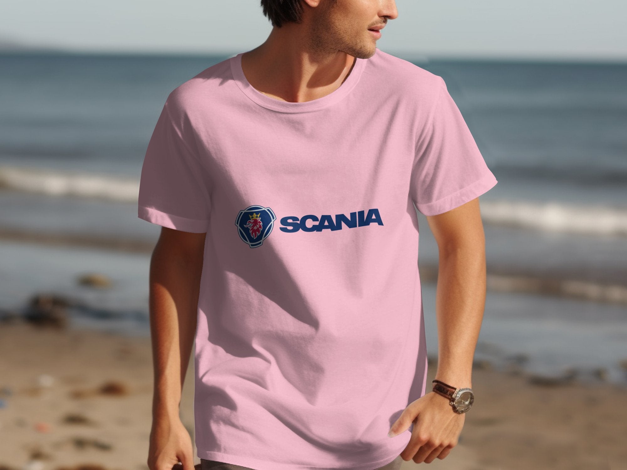 Dressed in a pink Scania logo T-shirt from Garment Graphics, a person stands by the shoreline. The calm ocean and sandy beach make a serene backdrop as they look right, their watch visible on their wrist.