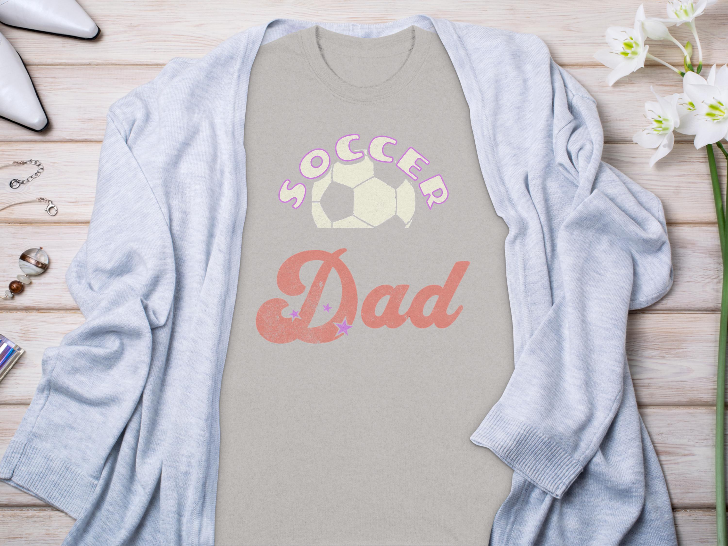 A Garment Graphics light gray Soccer Dad T-shirt with a soccer ball design is laid flat, perfectly paired with a cozy cardigan, white shoes, assorted jewelry, and a small plant on the wooden surface.