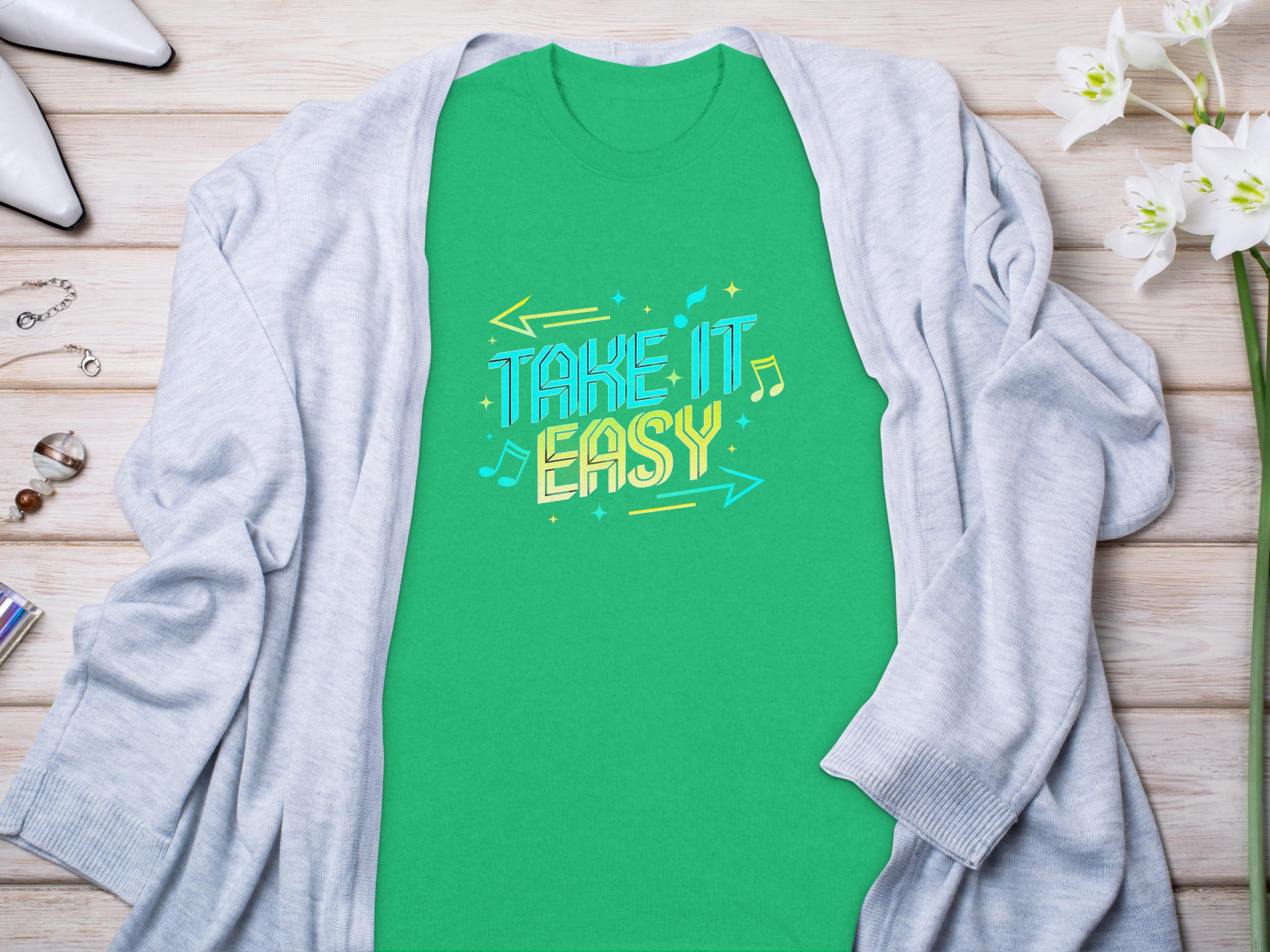 A vibrant Garment Graphics green Take It Easy T-Shirt with colorful letters, musical notes, and arrows lies flat on a wooden surface under a light gray cardigan, surrounded by white flowers, jewelry, and high-heeled shoes.