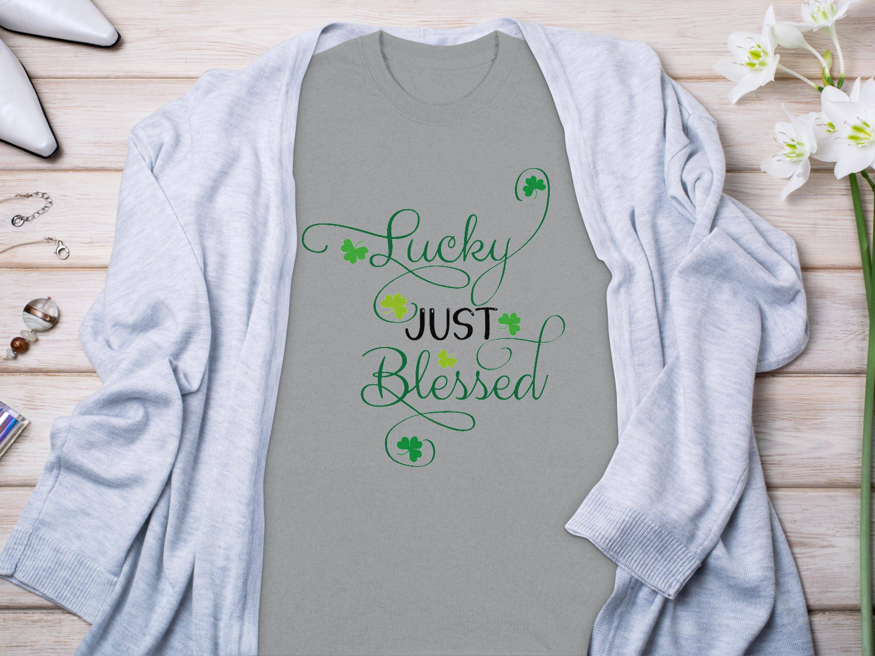 This Royal Mail - 24 Tracked classic fit gray t-shirt displays Lucky Just Blessed in green cursive with small shamrocks. Styled on a wooden surface, its paired with a gray cardigan, flowers, jewelry, and white shoes—an ideal addition to your graphic t-shirt collection.
