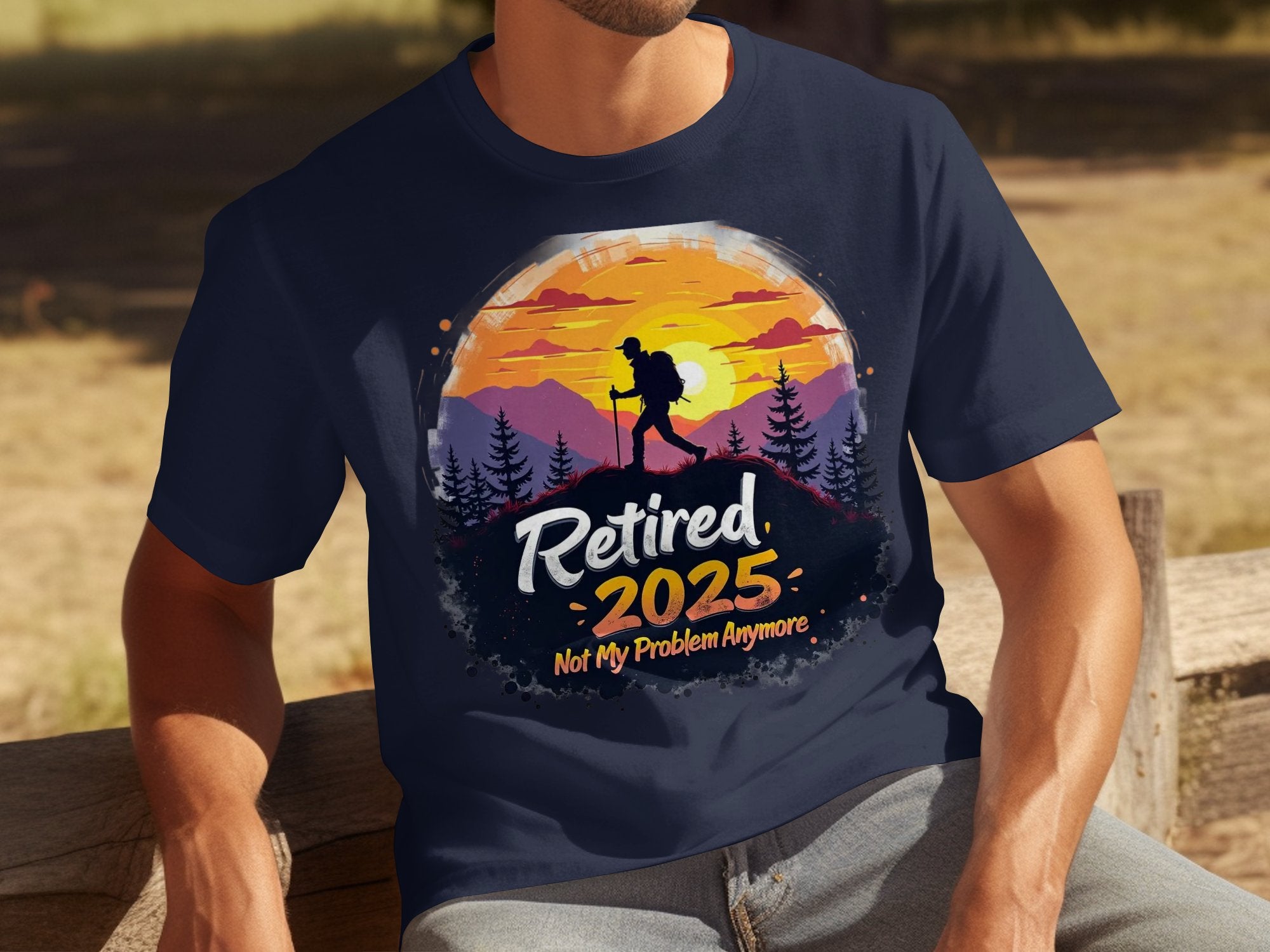 A person sits on a wooden bench wearing a navy T-shirt by Garment Graphics, depicting a hiker in front of a sunset with trees and the bold text Retired 2025: Not My Problem Anymore.