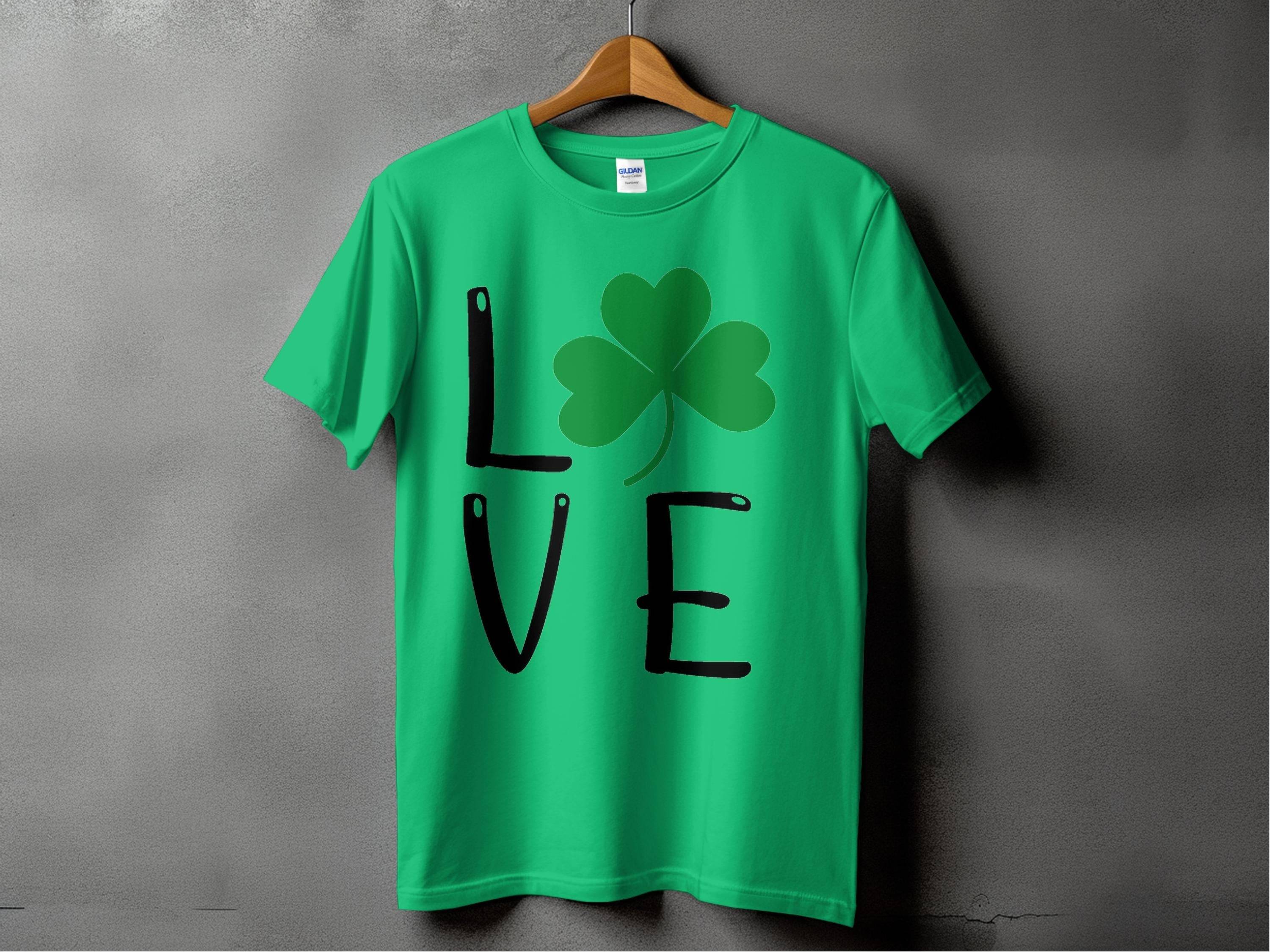 A green T-shirt on a hanger from Garment Graphics, featuring L♥VE with a shamrock replacing the O, embodies Irish spirit against a plain gray wall for St. Patricks Day.
