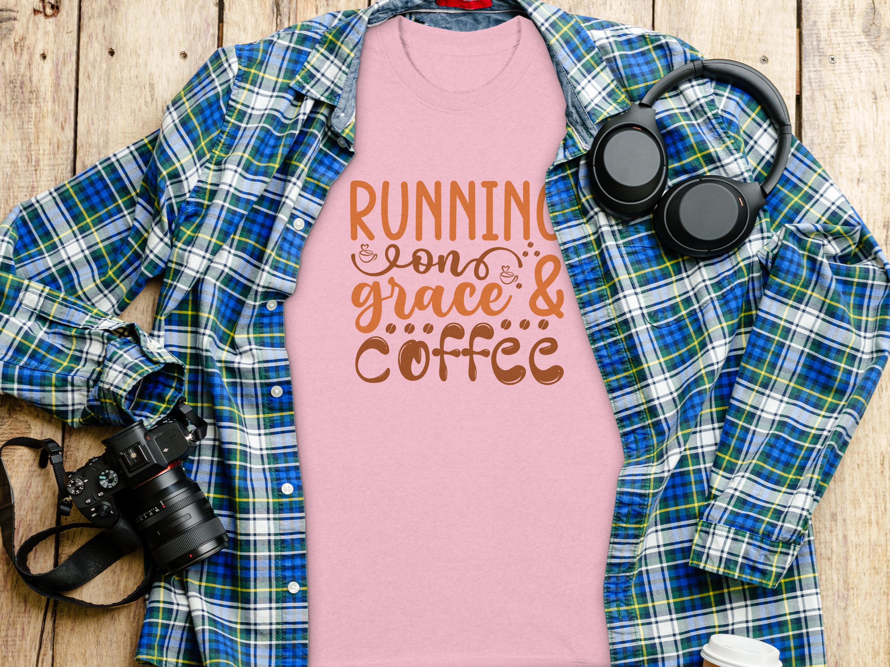 Garment Graphics pink Running on Grace & Coffee t-shirt is styled over a blue and green plaid shirt, set on a wooden surface. Accompanied by a black camera, headphones, and a white cup, this scene perfectly blends inspiration with style.