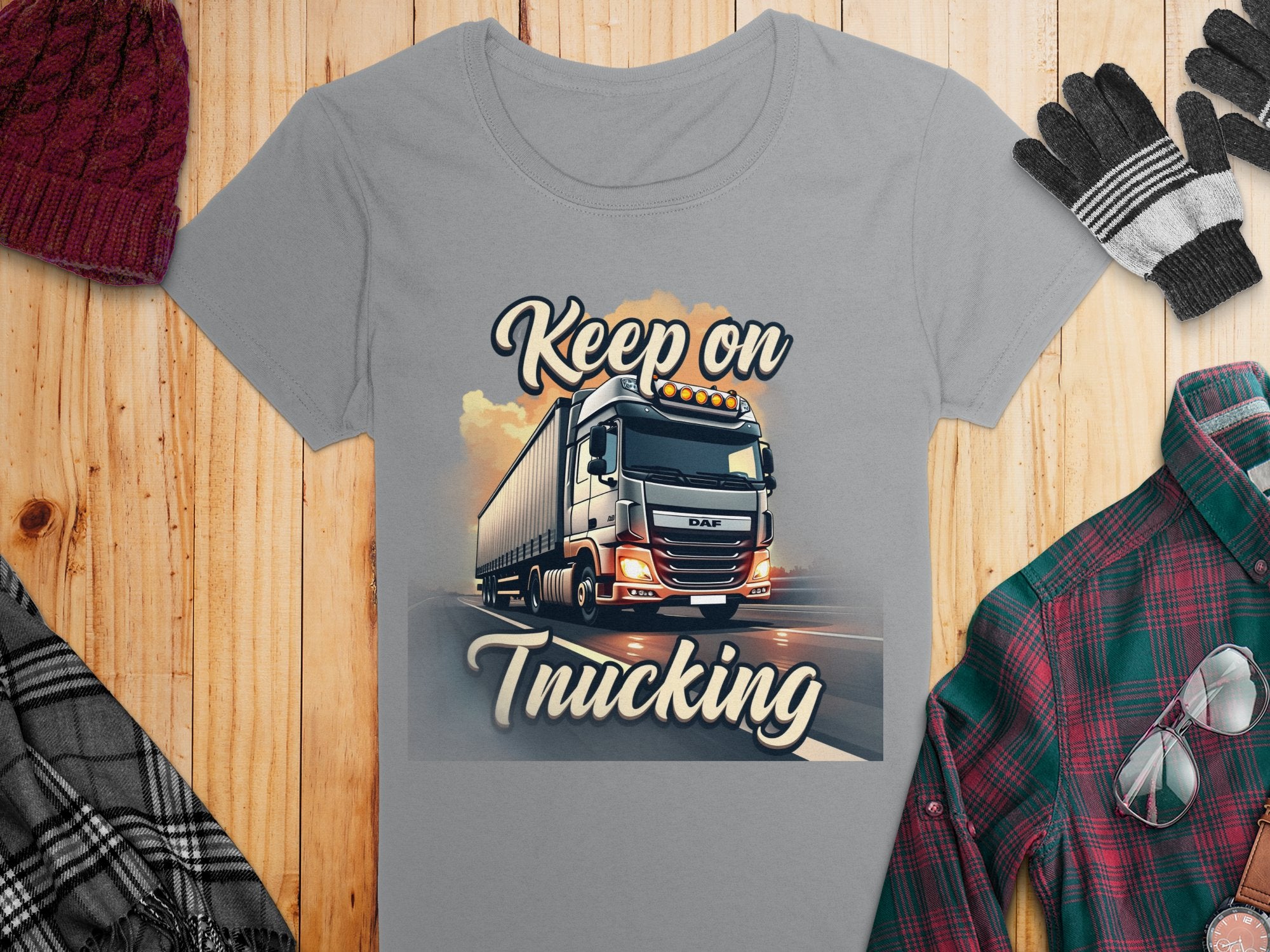A gray T-shirt by Garment Graphics lays on a wooden surface, featuring a truck graphic and Keep on Trucking text. Its surrounded by a maroon knit hat, gray gloves, and a green plaid shirt.