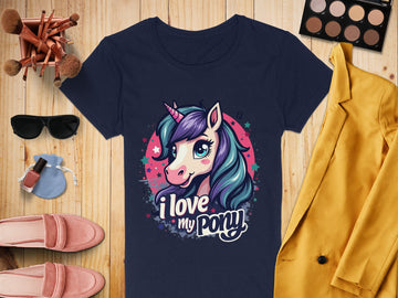 A Garment Graphics navy t-shirt with a whimsical unicorn graphic and I love my pony design rests on a wooden surface, perfect for unicorn lovers. Its surrounded by a yellow blazer, chic sunglasses, pink shoes, makeup essentials, nail polish, and a dainty purse.