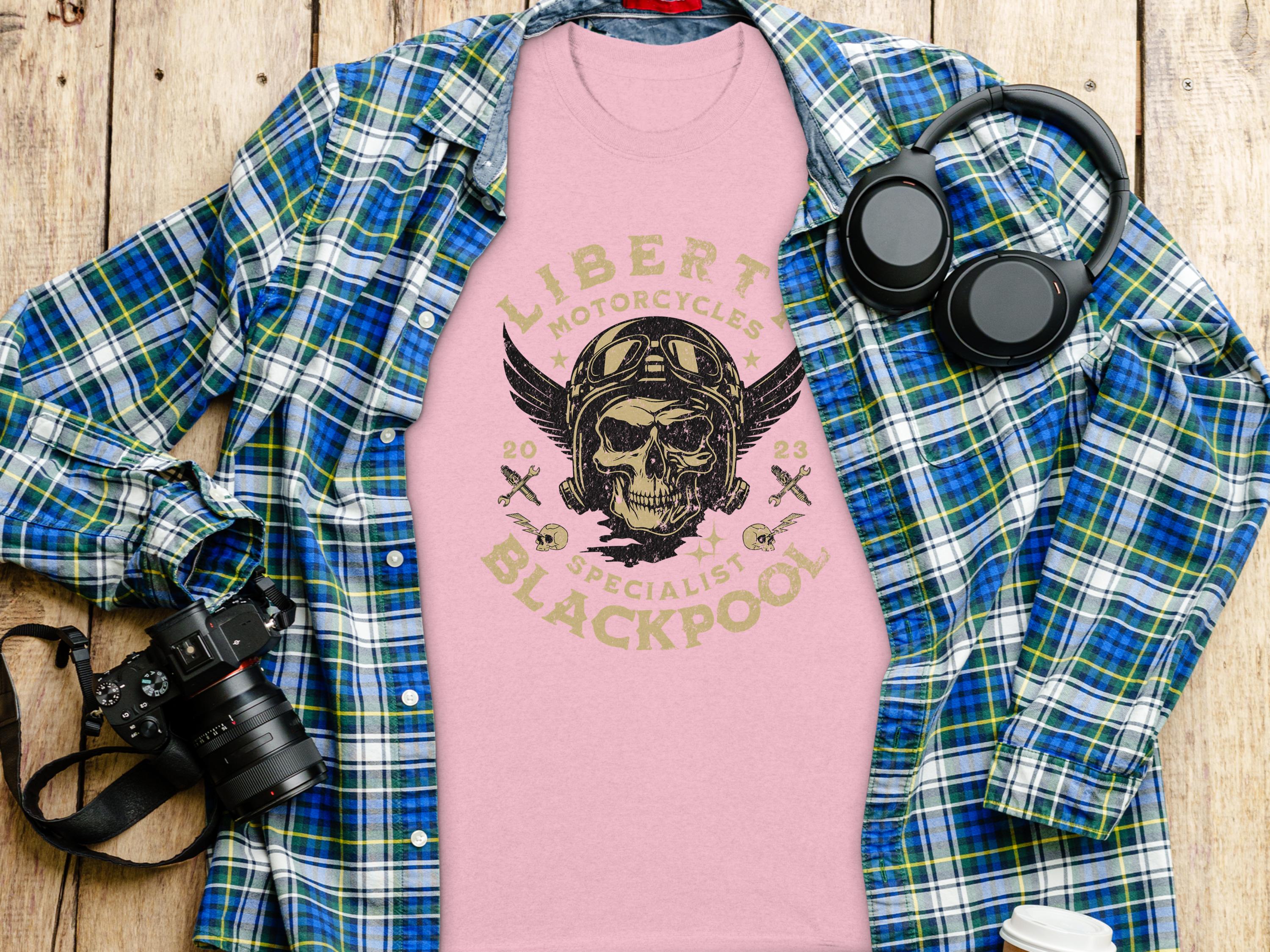 A flat lay showcases a pink Garment Graphics t-shirt with a skull and Liberty Motorcycles Blackpool 2023 Specialists print. Perfect for vintage design lovers and motorcycle enthusiasts, its surrounded by a blue-green plaid shirt, camera, and black headphones on a wooden surface.