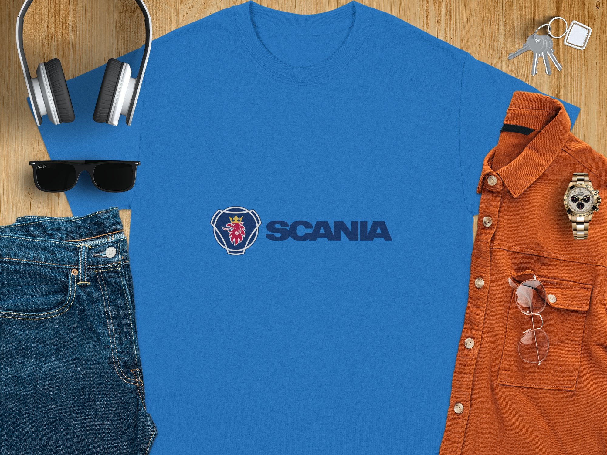 On a wooden surface, theres a blue Garment Graphics Scania Logo T-shirt, headphones, sunglasses, jeans, a brown button-up shirt, a watch, keys, and a smartphone. This neatly arranged flat lay highlights stylish Scania merchandise.