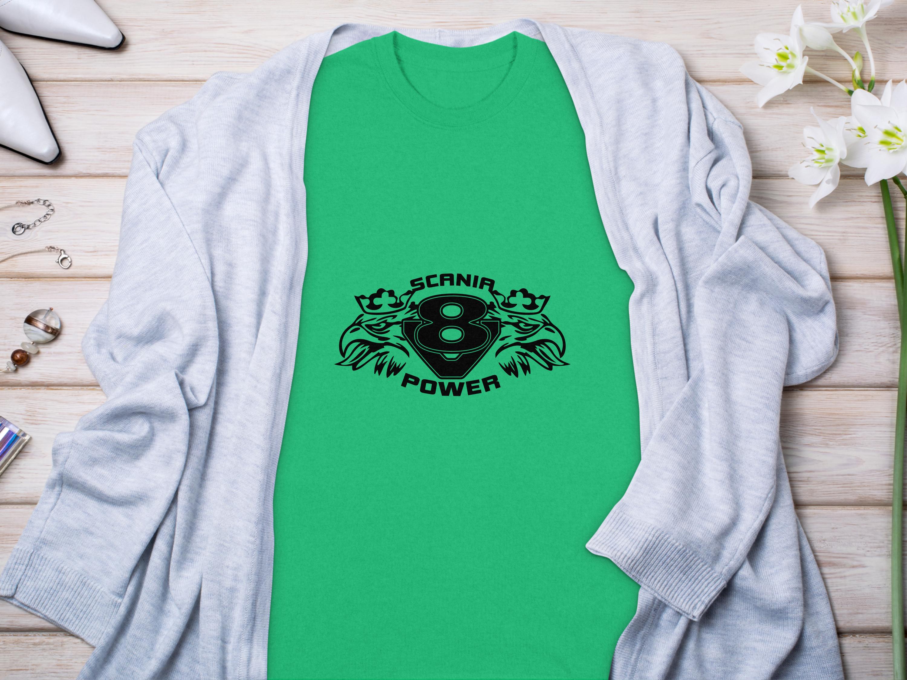 A green T-shirt featuring a black Scania 8 Power logo and two griffins by Garment Graphics lies flat, highlighting sleek design. Its paired with a light gray cardigan on wood, adorned with white orchids and jewelry.