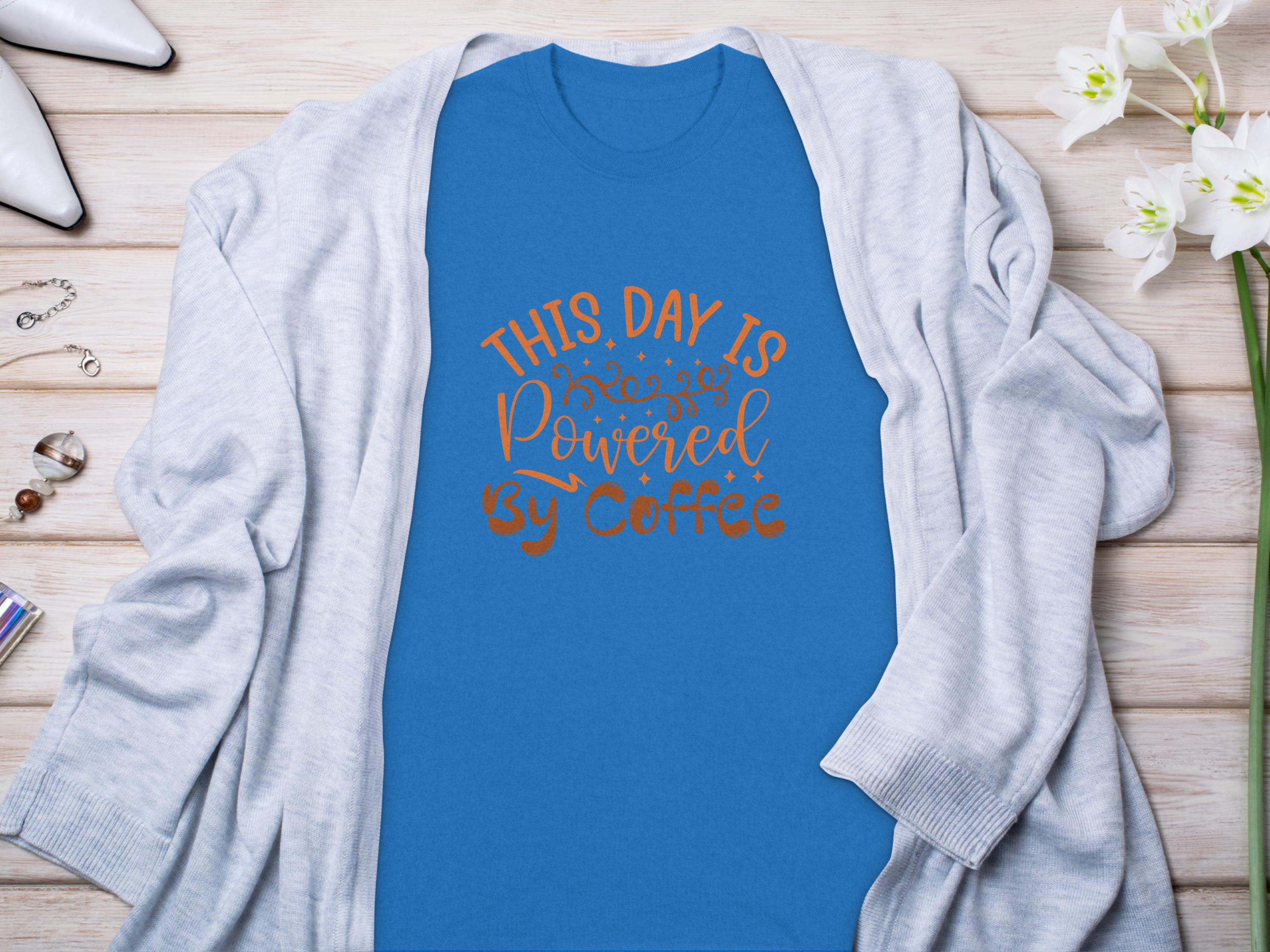 Garment Graphics classic blue T-shirt features This day is powered by coffee in decorative brown lettering. Styled flat with a light gray cardigan, white shoes, jewelry, and white flowers on a wooden surface, it embodies a positive vibe.