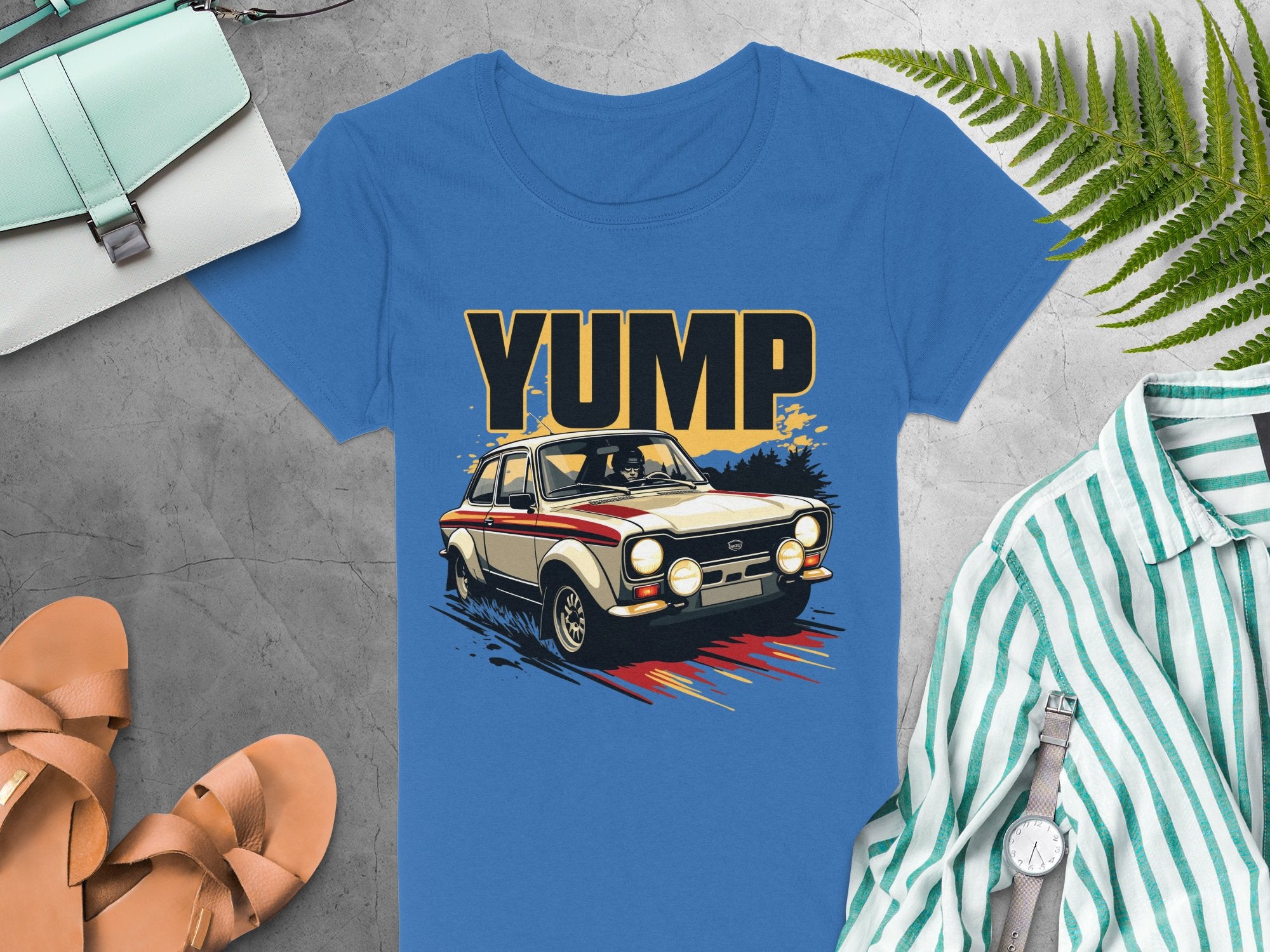 A bold graphic vintage car with YUMP is featured on this blue t-shirt by Garment Graphics. It is displayed on a gray background, accompanied by a striped shirt, brown sandals, light blue handbag, watch, and green fern leaves.