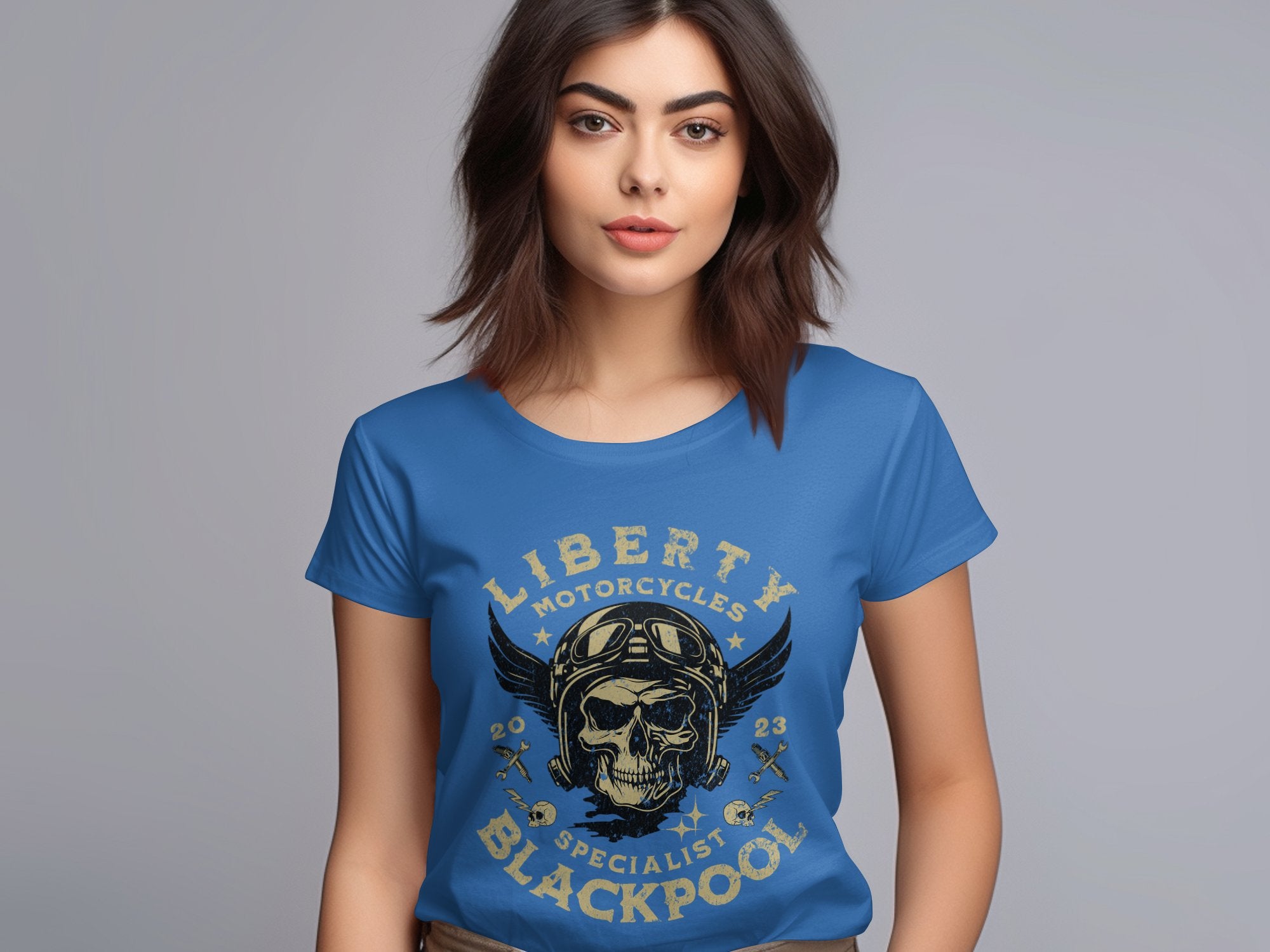 A woman with long dark hair wears a blue Garment Graphics t-shirt featuring Blackpool-themed design elements like a skull and wings motif. The text Liberty Motorcycles Specialist Blackpool is displayed as she stands against a plain gray background.