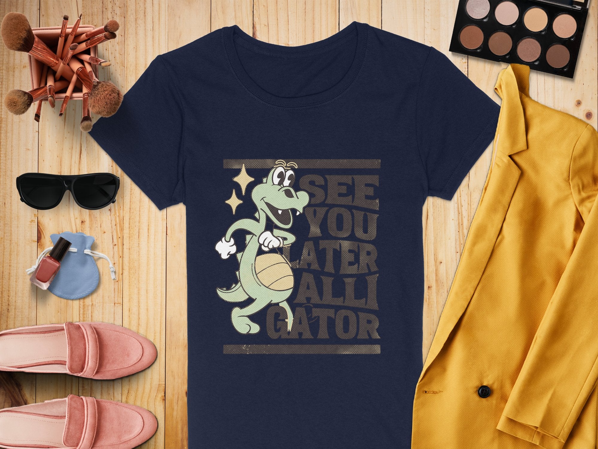 Garment Graphics navy blue alligator T-shirt features a humorous cartoon and See You Later Alligator text, styled for women with a yellow blazer, makeup palette, brushes, sunglasses, pink loafers, a small pouch, and nail polish on a wooden surface.