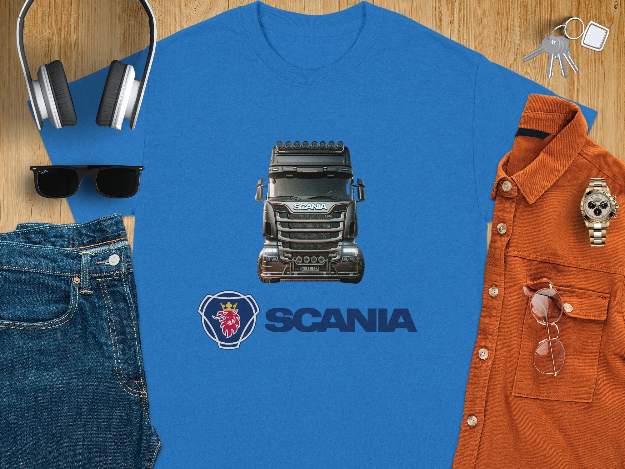 Displayed on a wooden surface, this blue T-shirt from Garment Graphics is ideal for truck lovers, showcasing a bold Scania truck design with the Scania logo below. Its styled with jeans, sunglasses, headphones, keys, a brown shirt, and a watch for the ultimate trendy look.