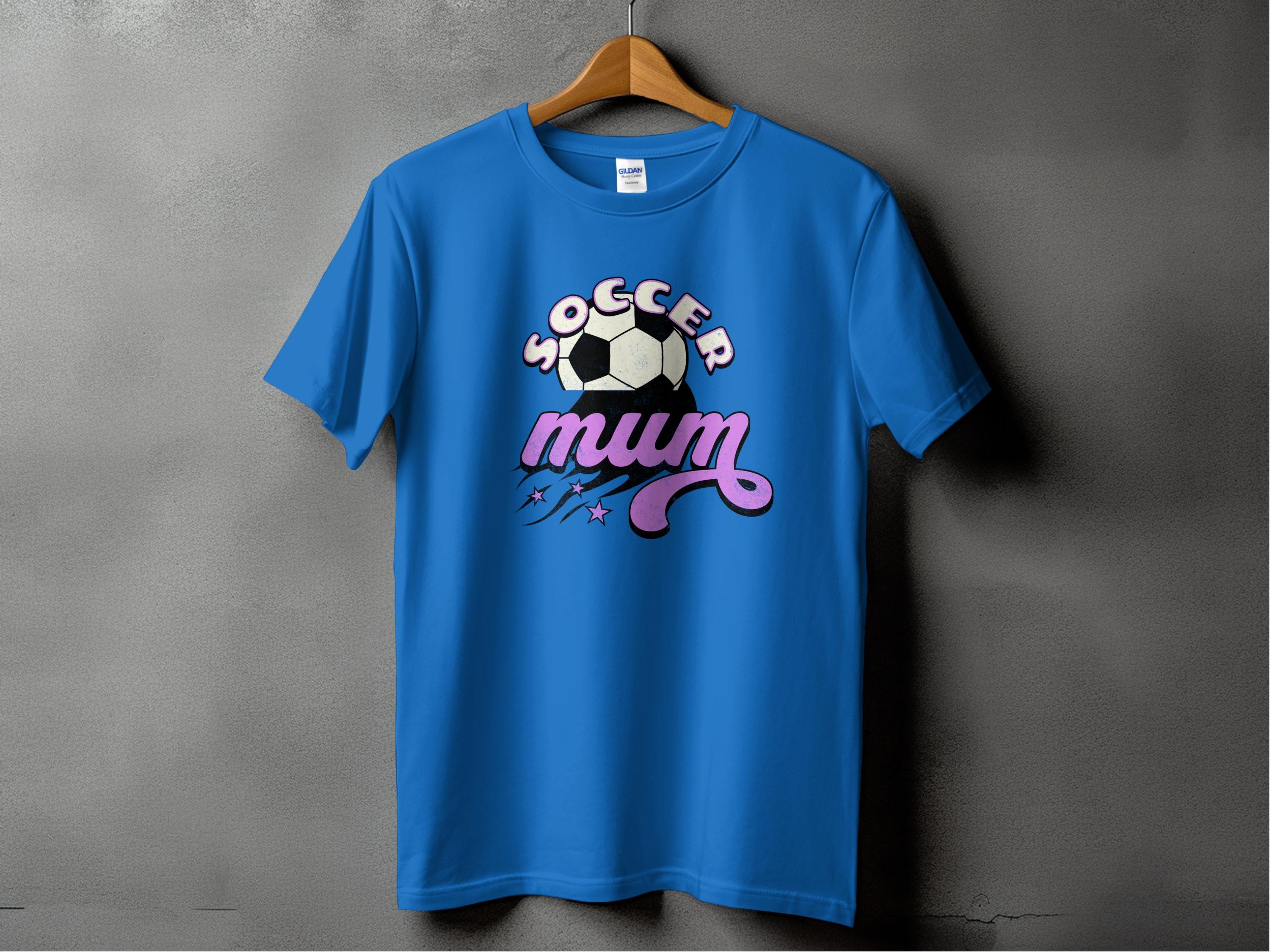 A Garment Graphics classic fit blue T-shirt hangs on a wooden hanger against a gray wall, featuring a soccer ball graphic with Soccer Mum in bold letters and vibrant purple and pink star and swirl accents.